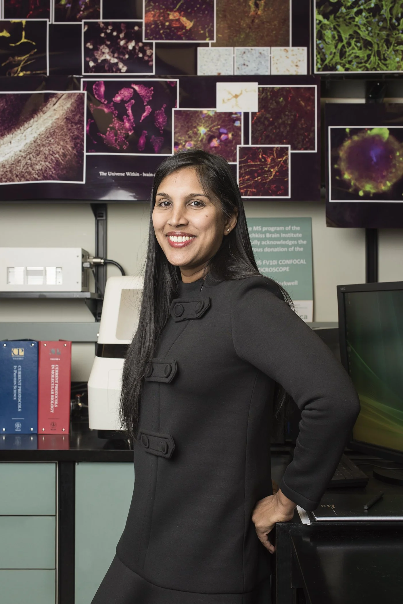 A woman with long dark hair smiling in a laboratory or research environment, with a wall of colorful microscopic images and computer monitors behind her.