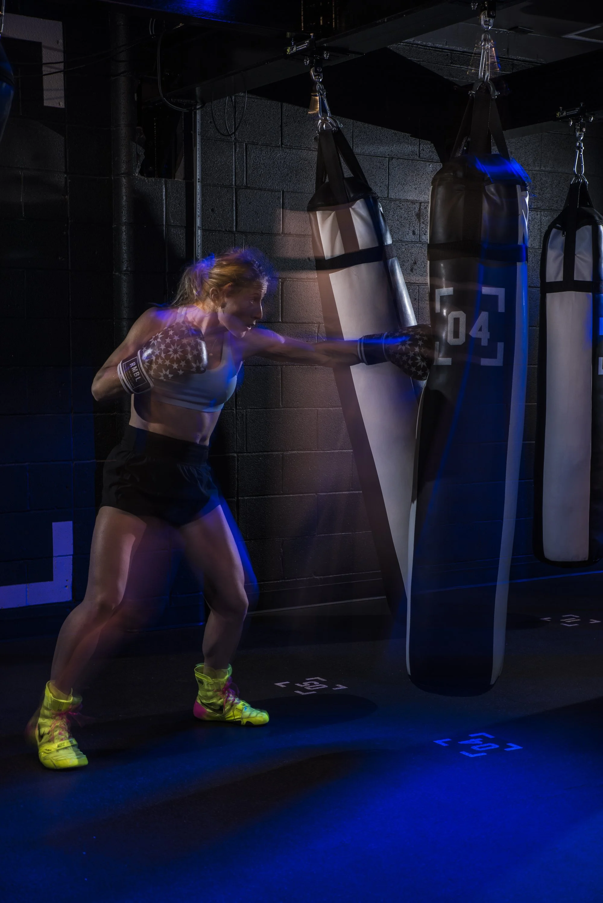 A woman wearing boxing gloves and athletic attire punches a hanging punching bag in a dark gym lit by colored lights.