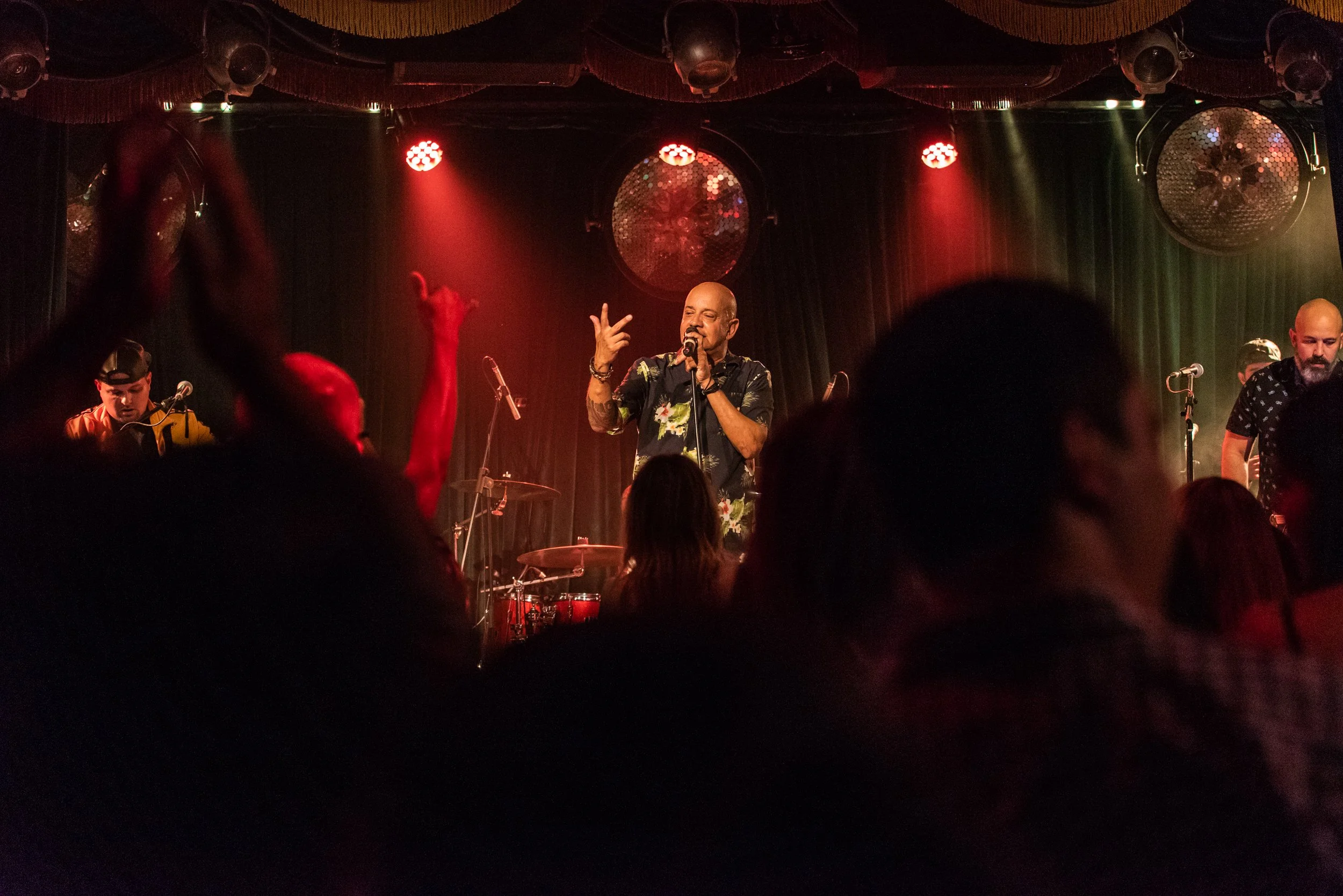 Performer singing on stage with a microphone, accompanied by band members, in a dimly lit venue with red and green lighting.