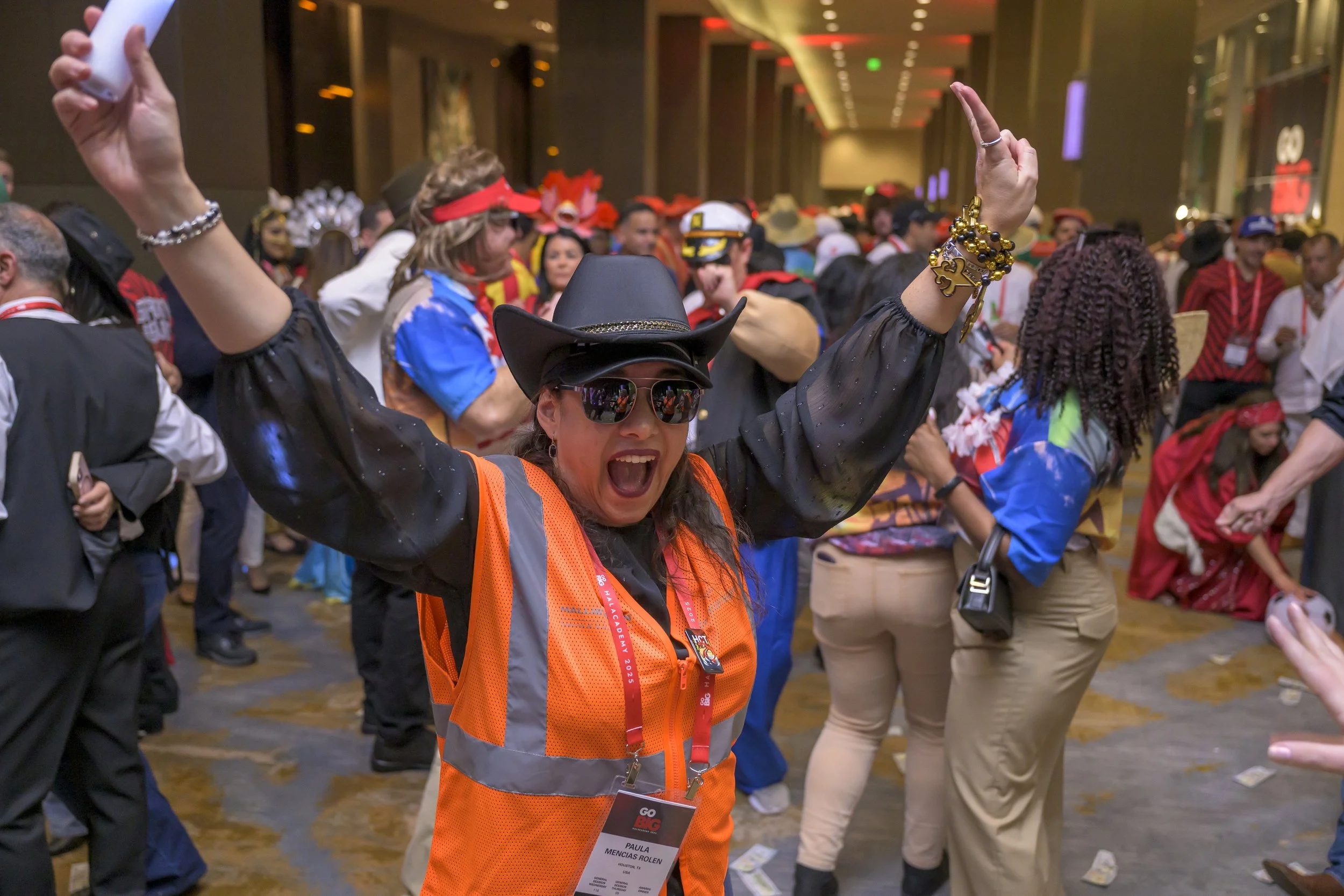 A woman at a lively indoor party, wearing a cowboy hat, sunglasses, and an orange safety vest, with her arms raised and mouth open in excitement, surrounded by people in colorful costumes and costumes, with confetti and money on the floor.
