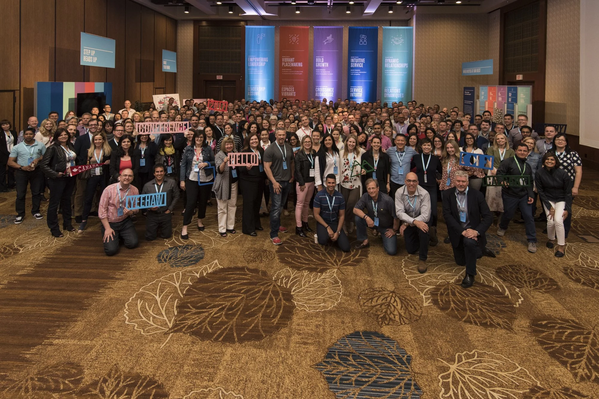 A large group of people gathered at a conference or event in a ballroom, holding signs and wearing name tags. The background has colorful banners with empowering messages.