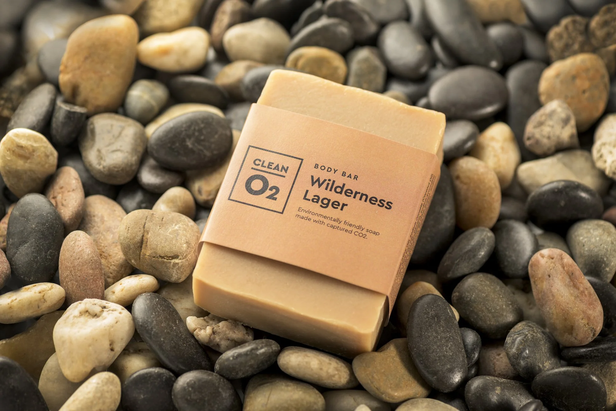 A bar of soap labeled Wilderness Lager from Clean O2 resting on a bed of multicolored small rocks.