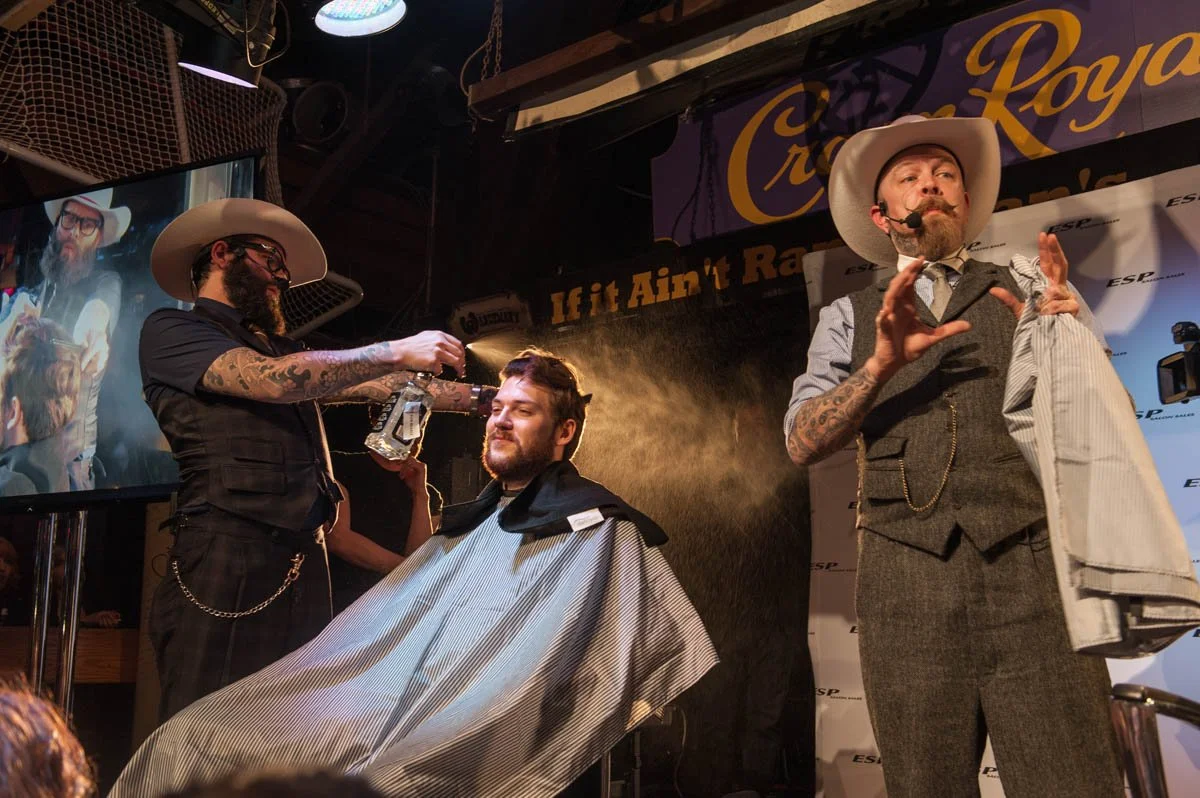 A man getting a haircut at a barbershop event, with two barbers in vintage-style attire, one of whom is holding a spray bottle. The man is smiling and wearing a barber cape. The background features a TV screen and banners for a men's grooming event.