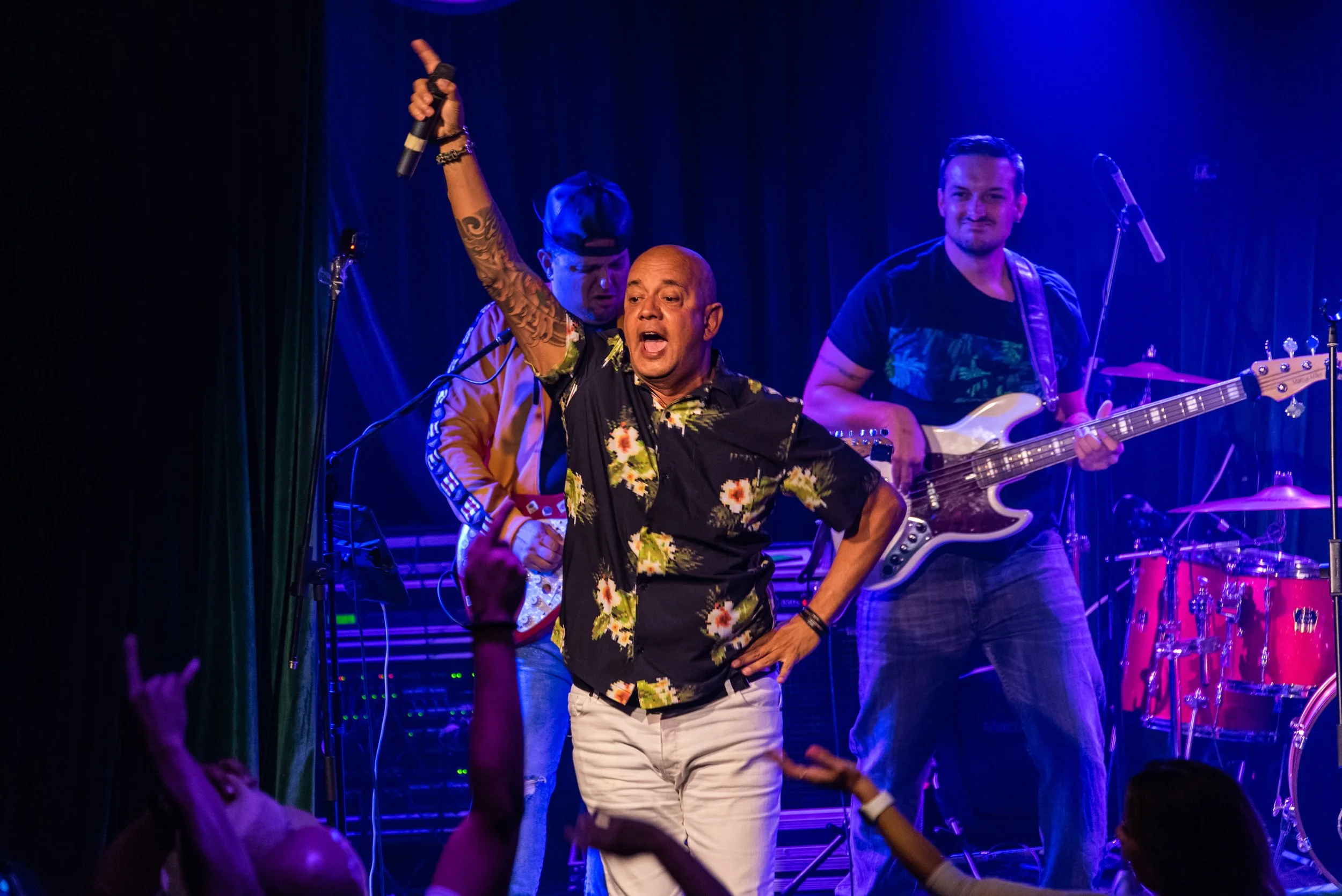 A lively band performs on stage with a singer holding a microphone, wearing a floral shirt, and audience members raising their hands.