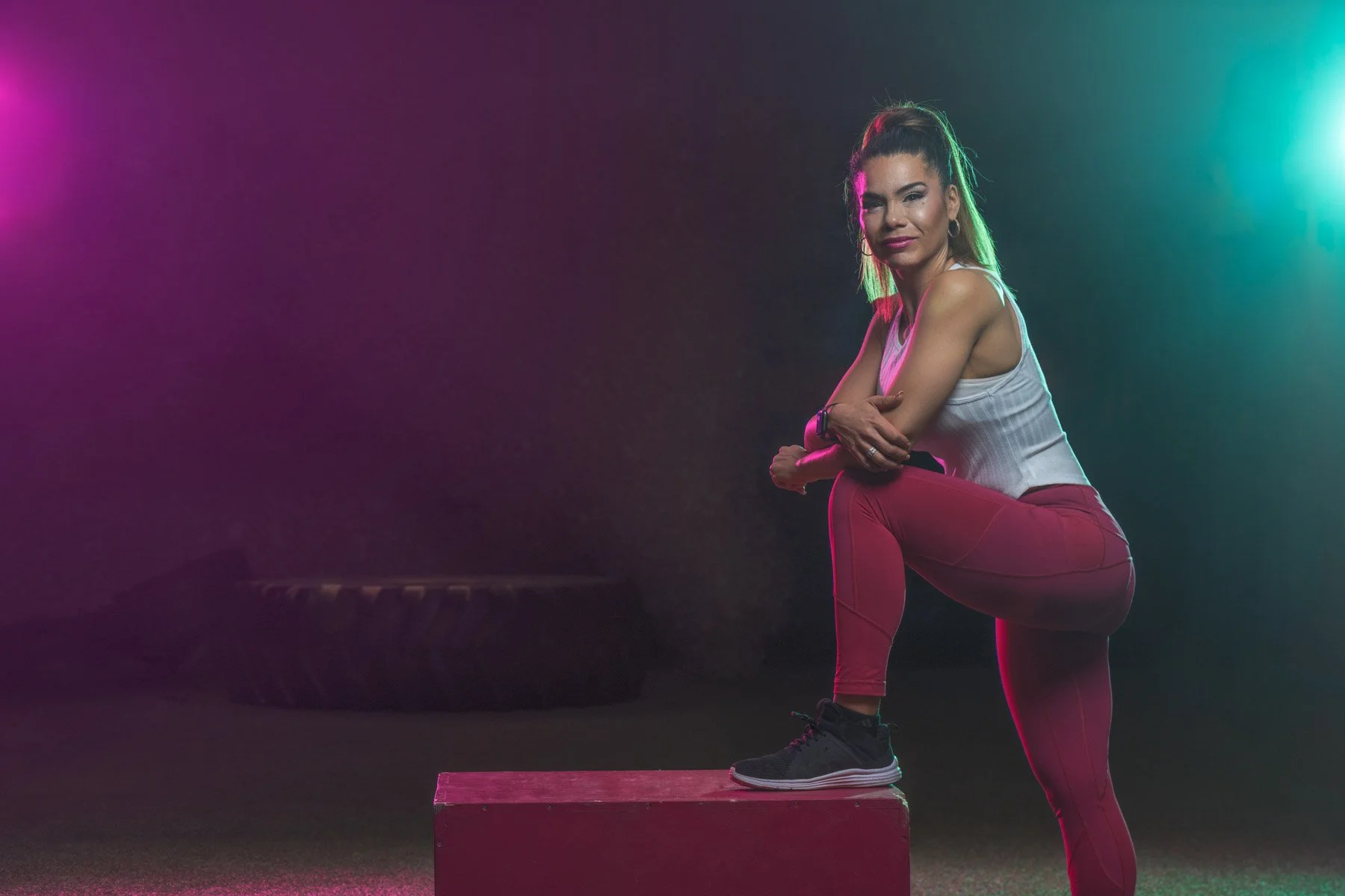 A woman in workout clothes, standing with one foot on a pink box, in a gym with colorful lighting.