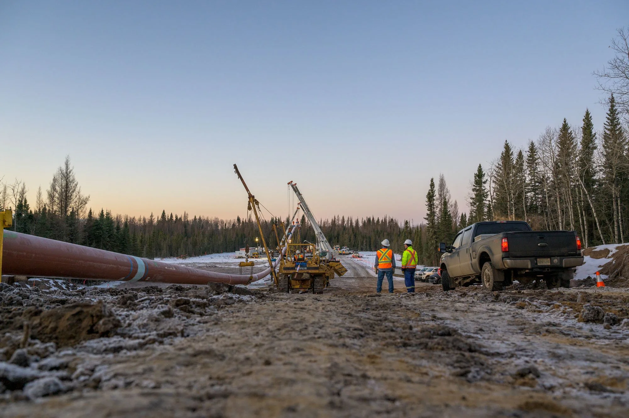 Construction workers in safety vests and helmets working on a pipeline in a snowy forest area during late afternoon or early evening.
