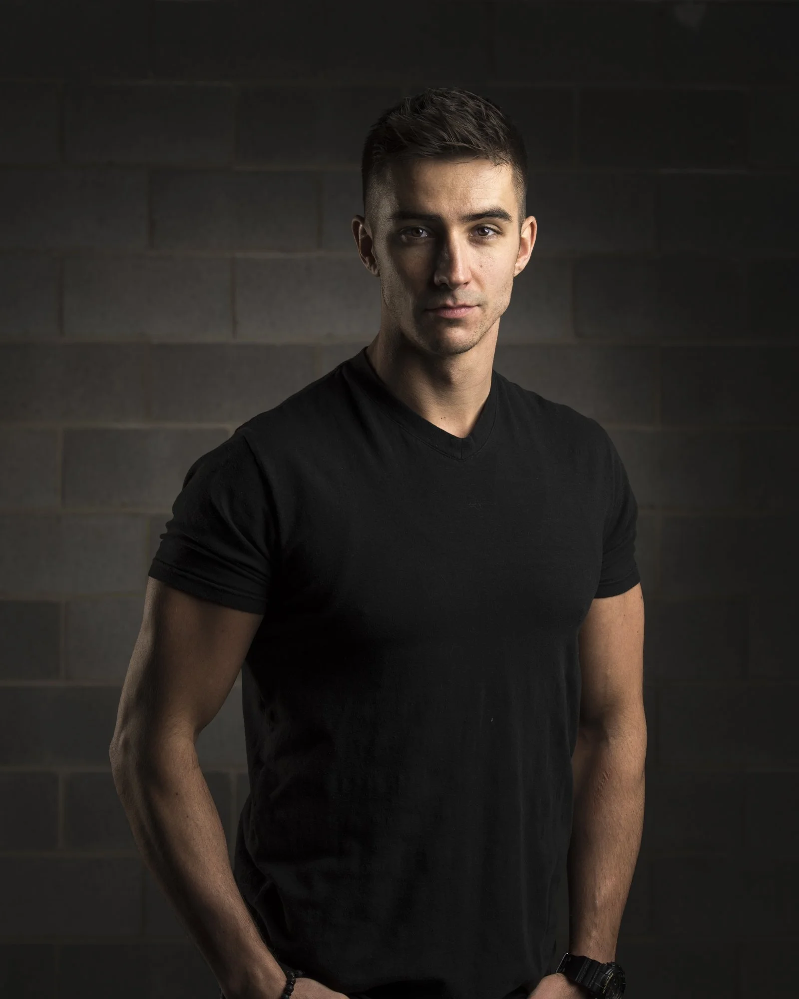 A young man with short dark hair, wearing a black V-neck T-shirt, standing against a dark gray brick wall, looking directly at the camera.