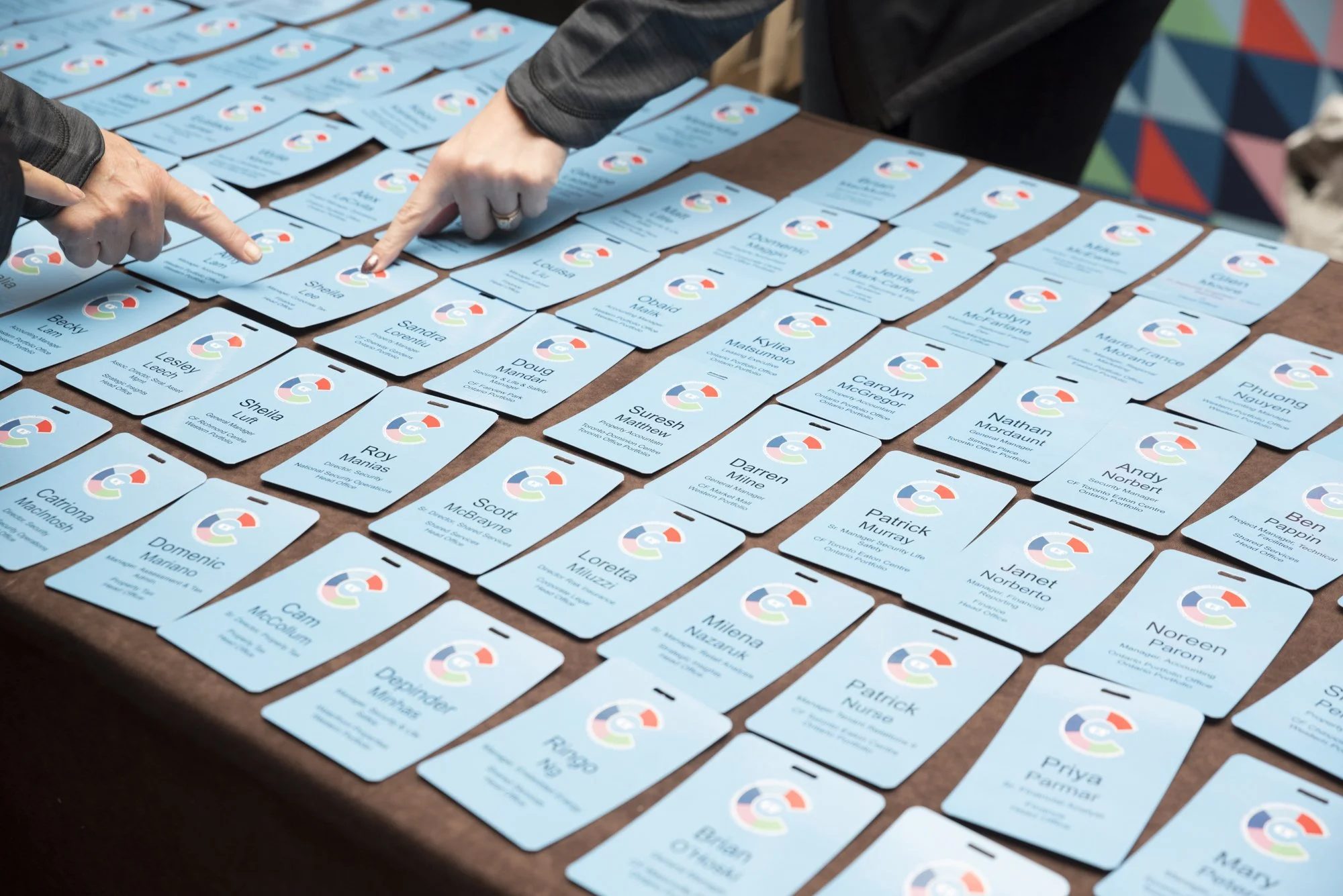 Multiple light blue conference badges laid out on a table with two people pointing at them.