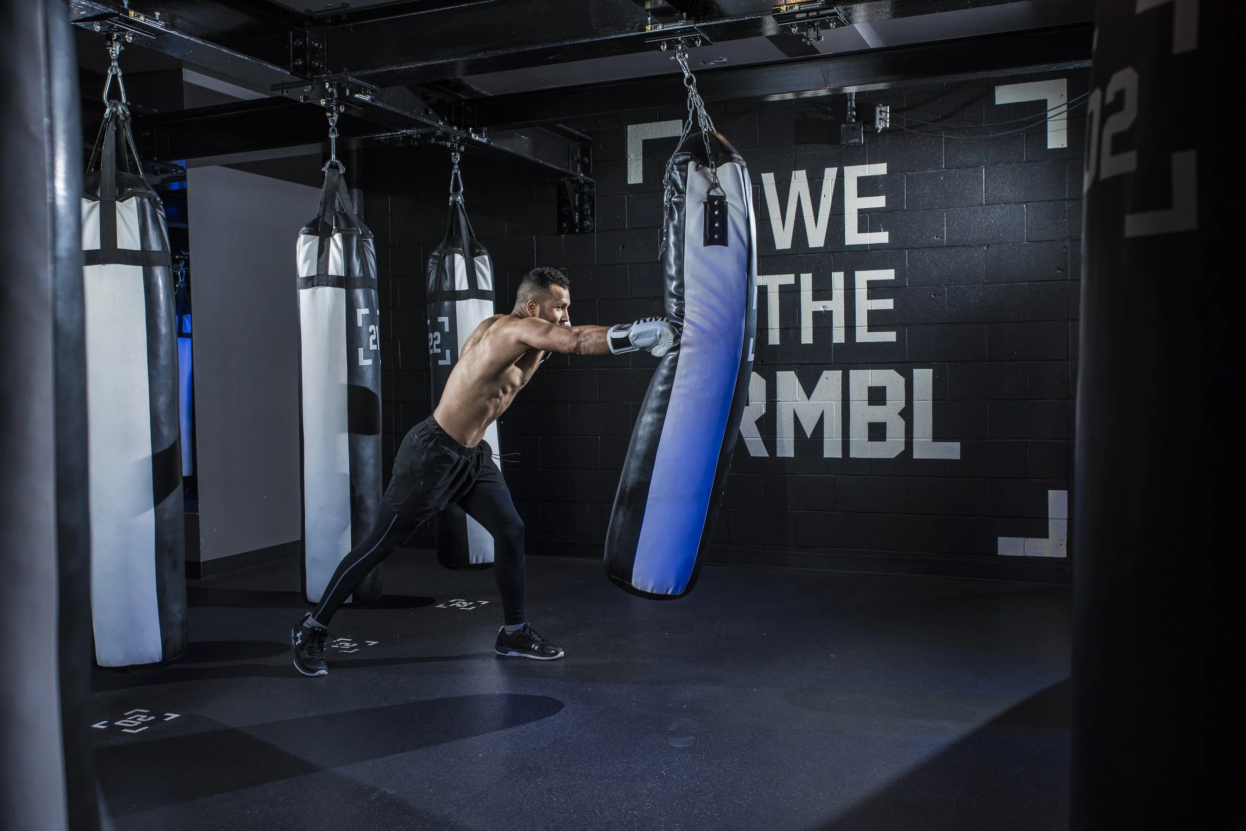 A shirtless man wearing black athletic wear and gloves is punching a blue and black heavy bag in a gym with black brick walls and several hanging other punching bags.