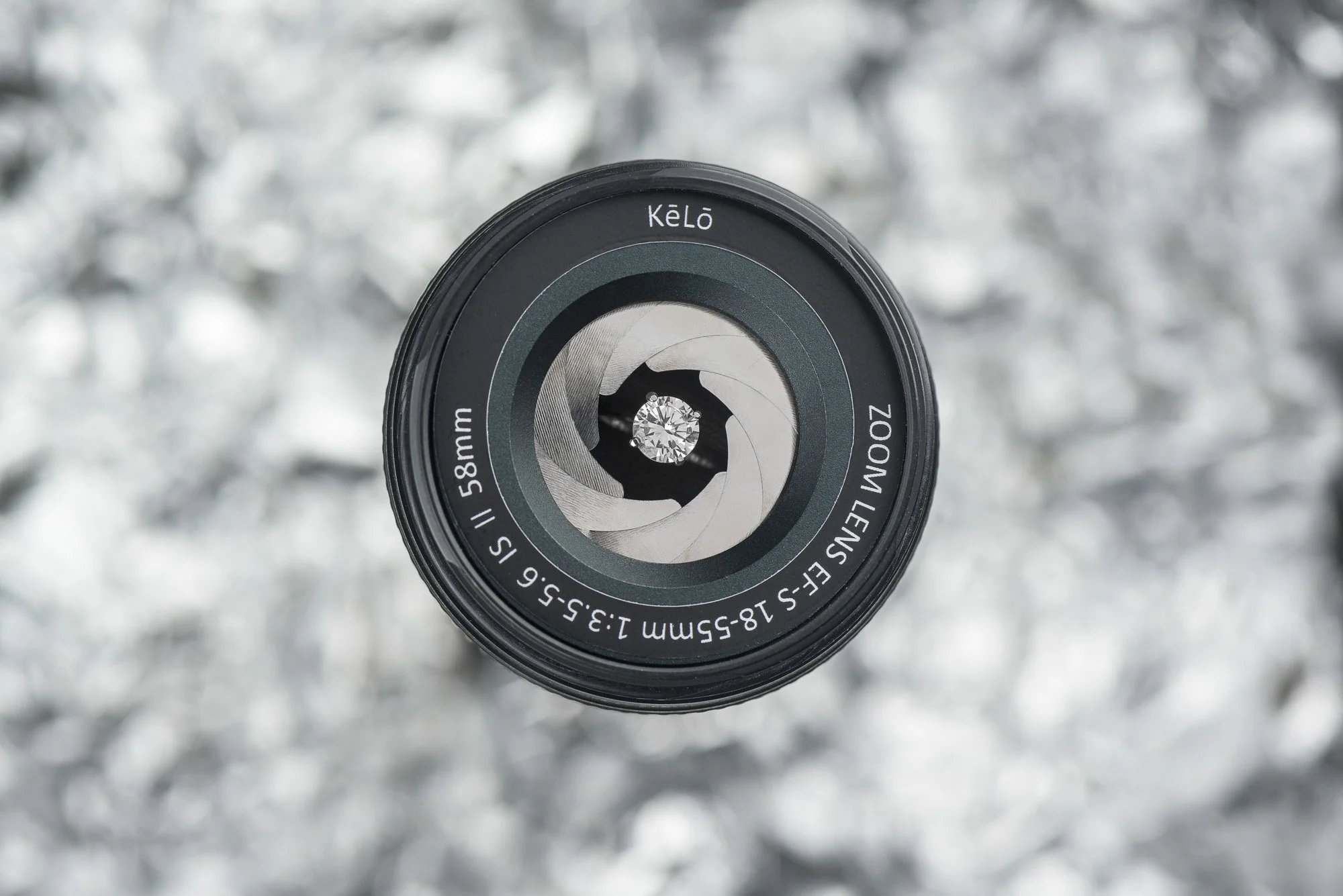 Camera lens with a diamond in the center, viewed from above, against a blurred metallic background.