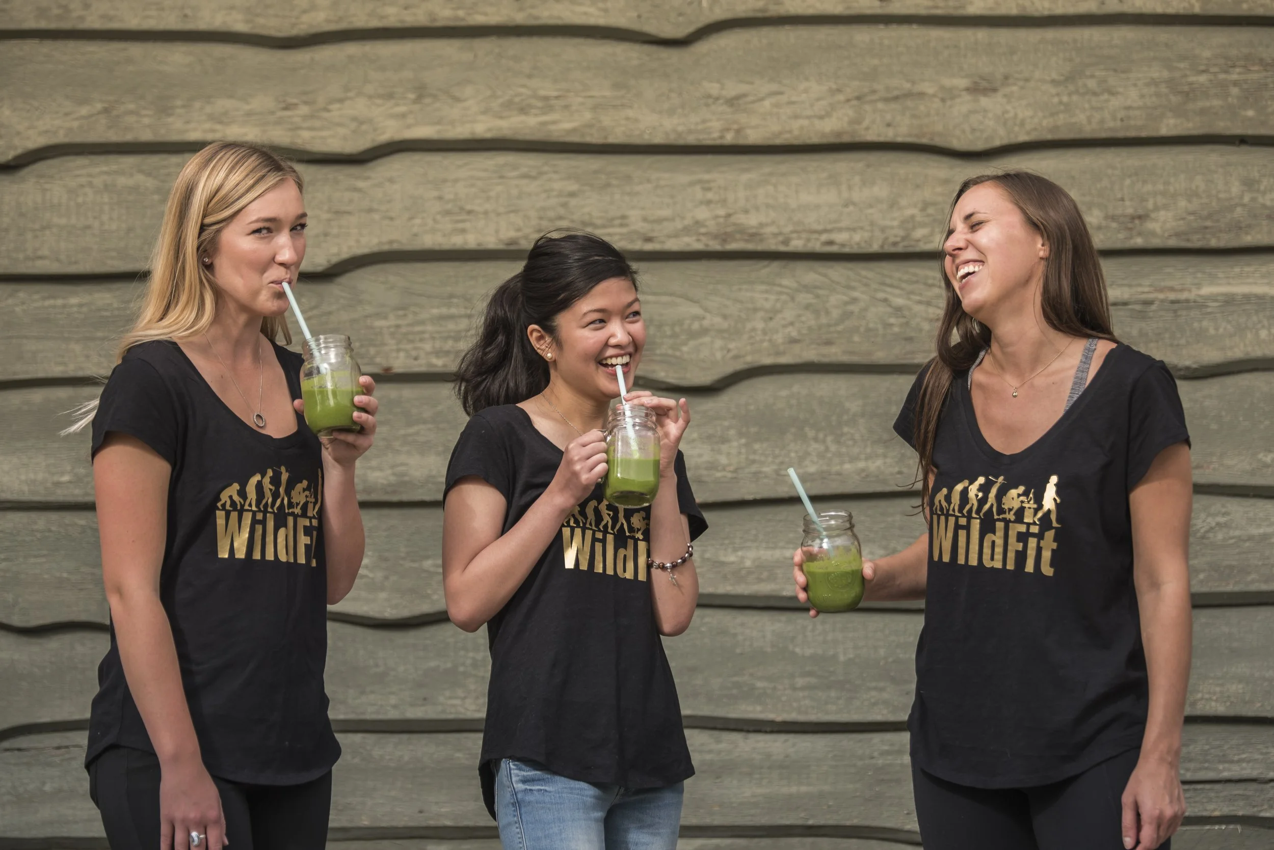 Three women wearing black 'WildFit' t-shirts stand against a wooden wall, each holding a green smoothie in a jar with a straw, sharing a cheerful moment together.