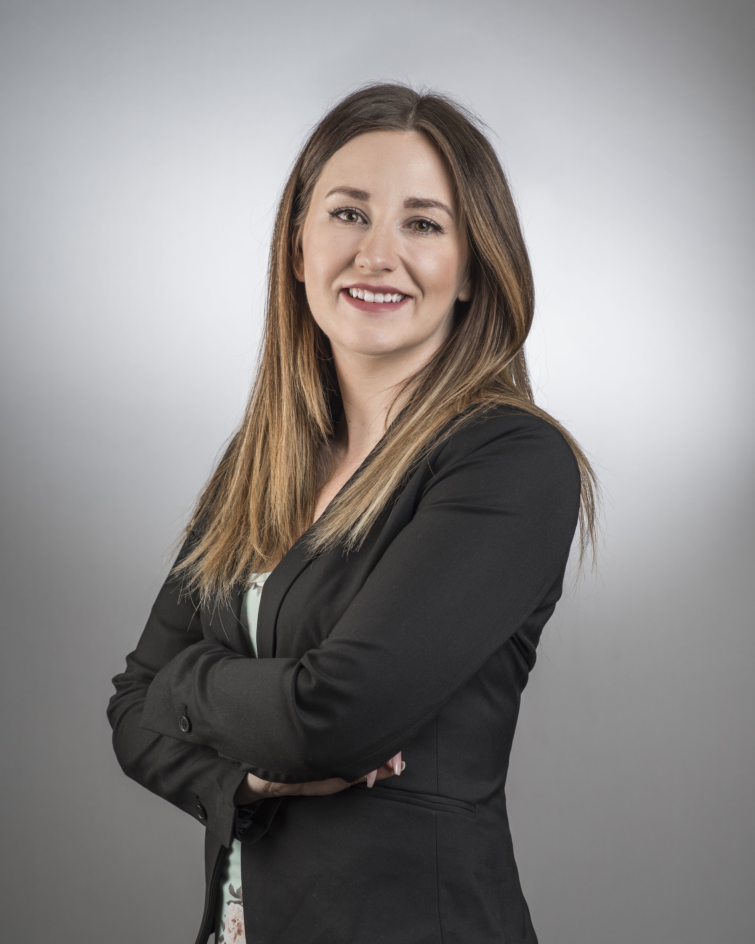 A professional woman with long brown hair and a black blazer smiling at the camera with arms crossed against a neutral background.