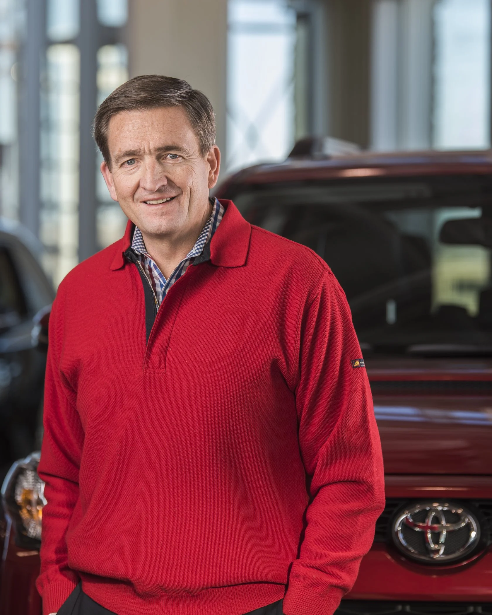 A man standing indoors next to a red Toyota vehicle, wearing a red sweater and a checkered shirt, smiling at the camera.