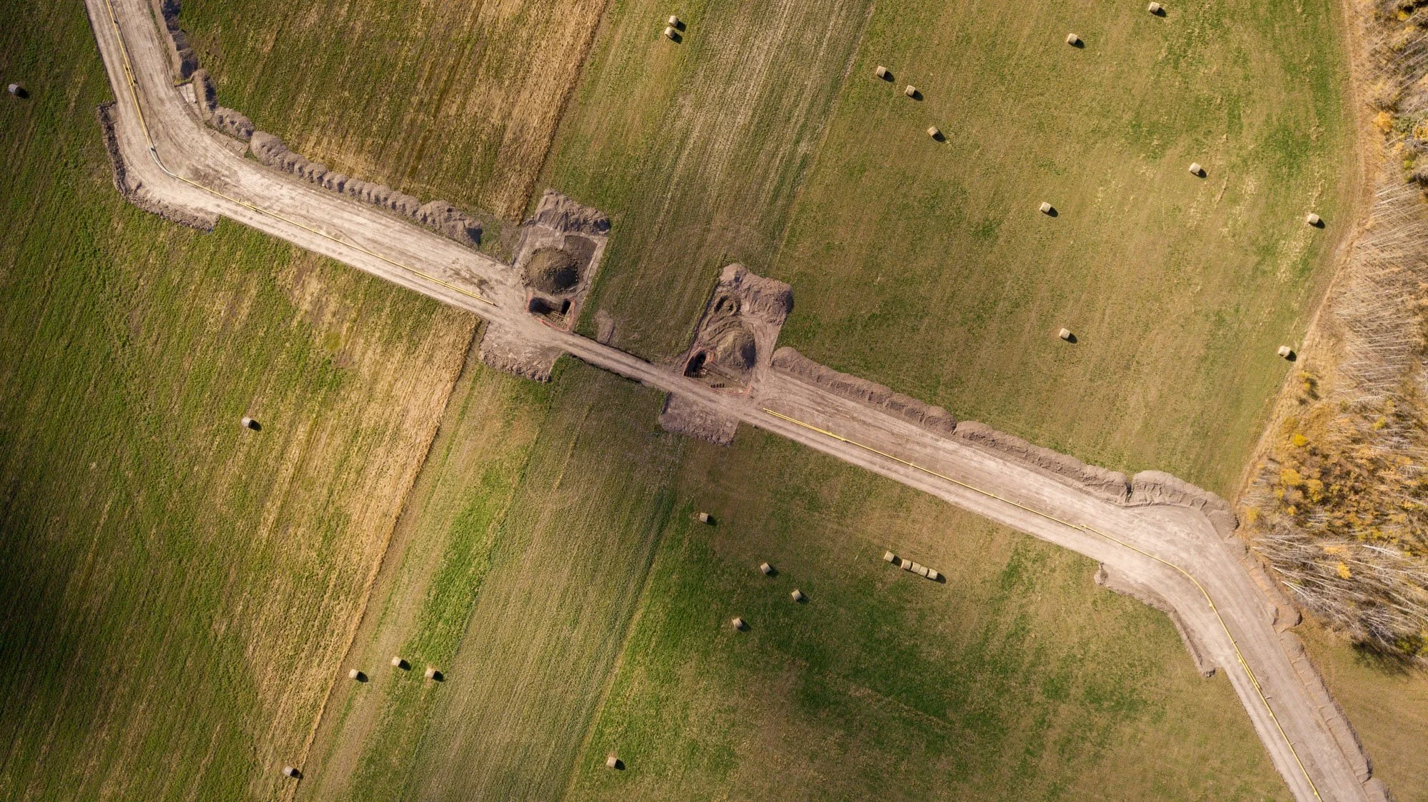 An aerial view of a farm field with hay bales scattered across the grass and a dirt pathway running through the center, flanked by trees on one side.
