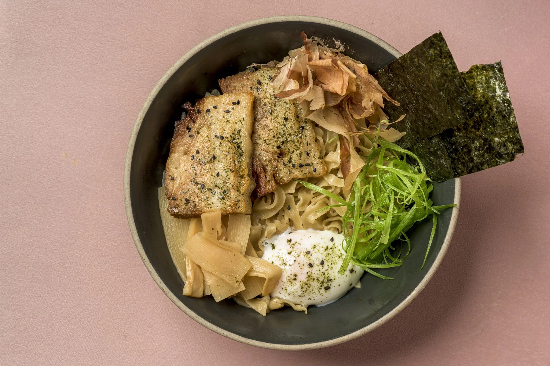 A bowl of ramen with grilled chicken, bamboo shoots, seaweed, green onions, a soft-boiled egg, and bonito flakes on a pink background.