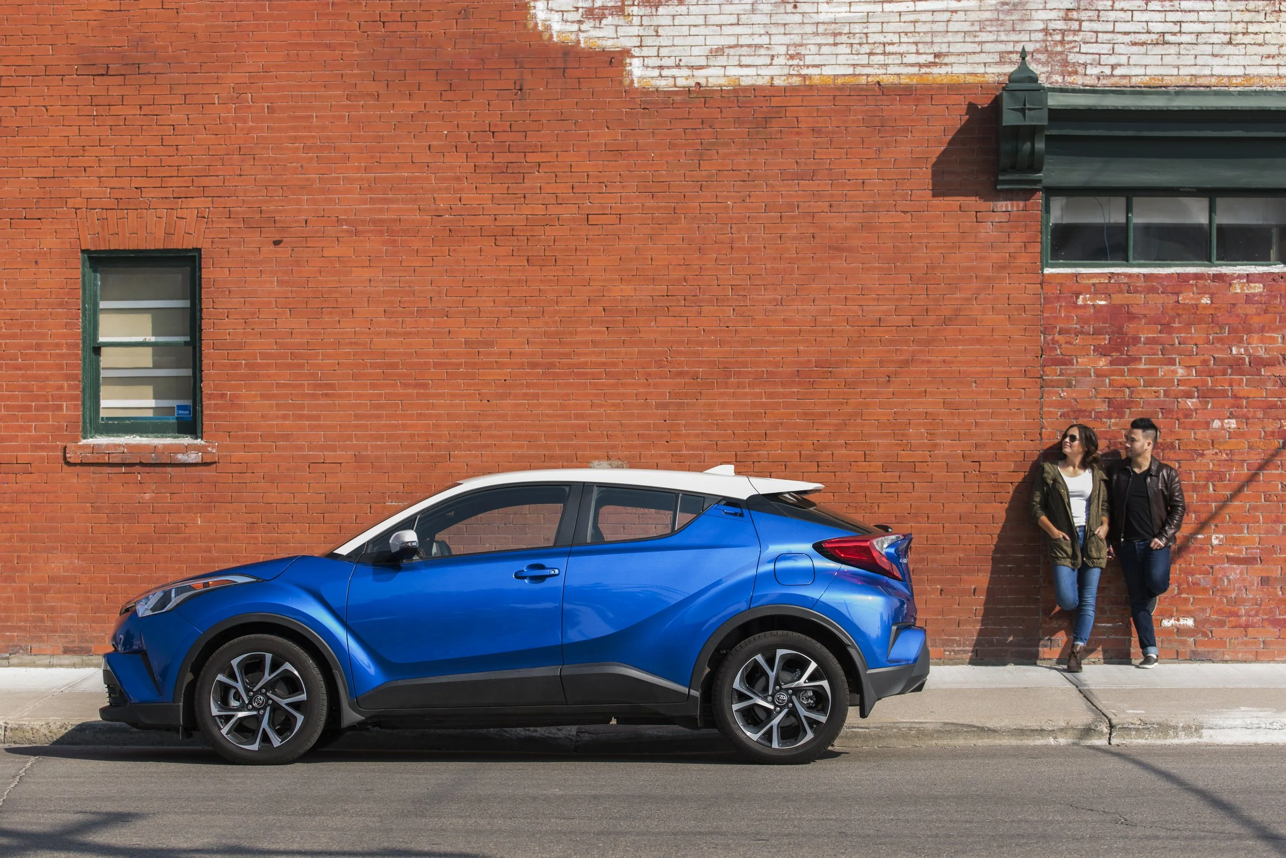 A blue compact SUV parked on the street beside a red brick wall, with a woman and a man standing and leaning against the wall, both dressed in casual attire.