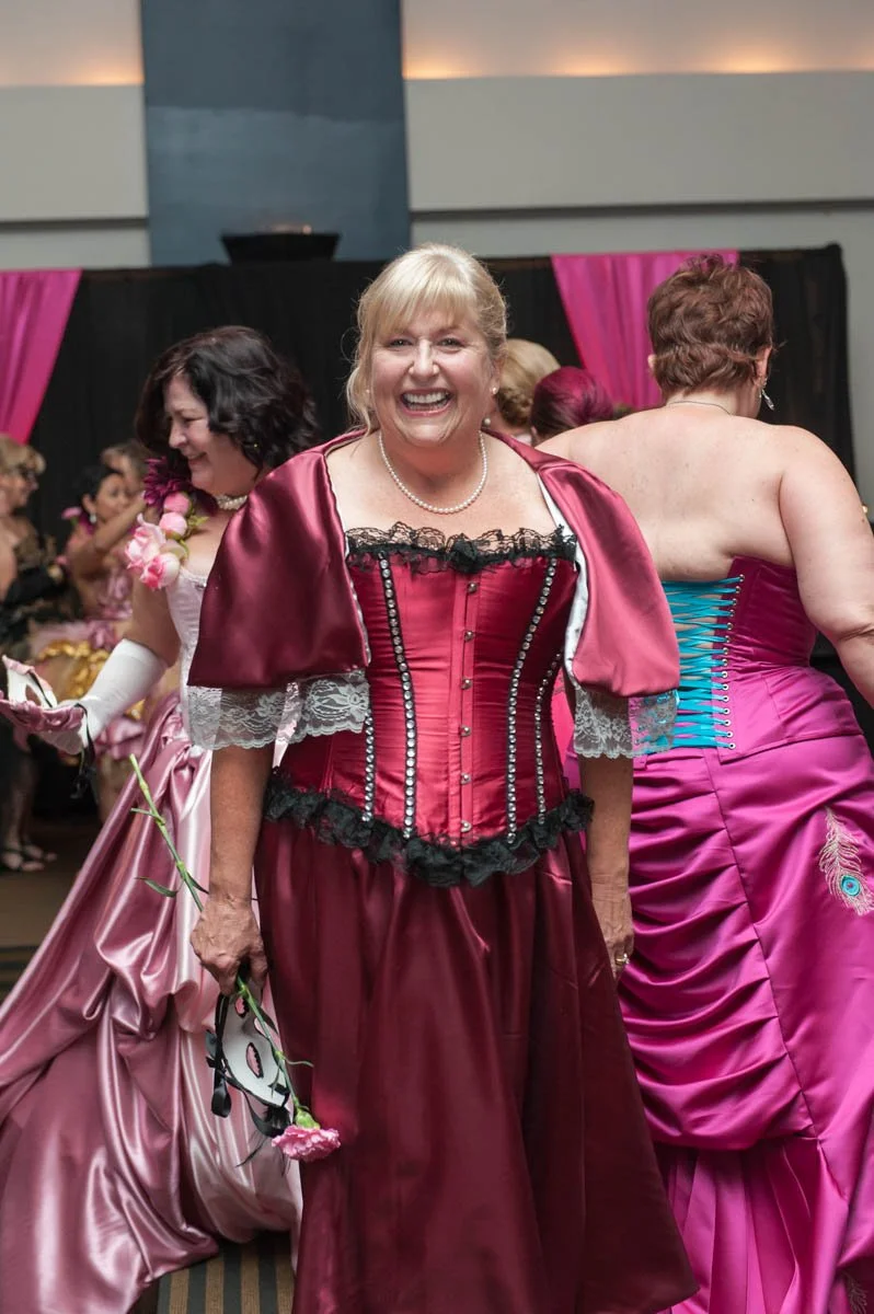 A group of women dressed in elaborate vintage-style dresses with corsets and satin fabrics, participating in a social event or dance in an indoor ballroom setting.