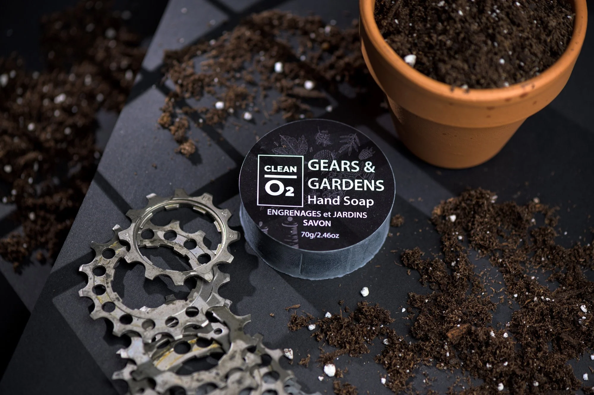 A roll of Gears & Gardens hand soap, a terracotta pot filled with soil, bicycle gears, dirt, and soil particles all placed on a black surface.