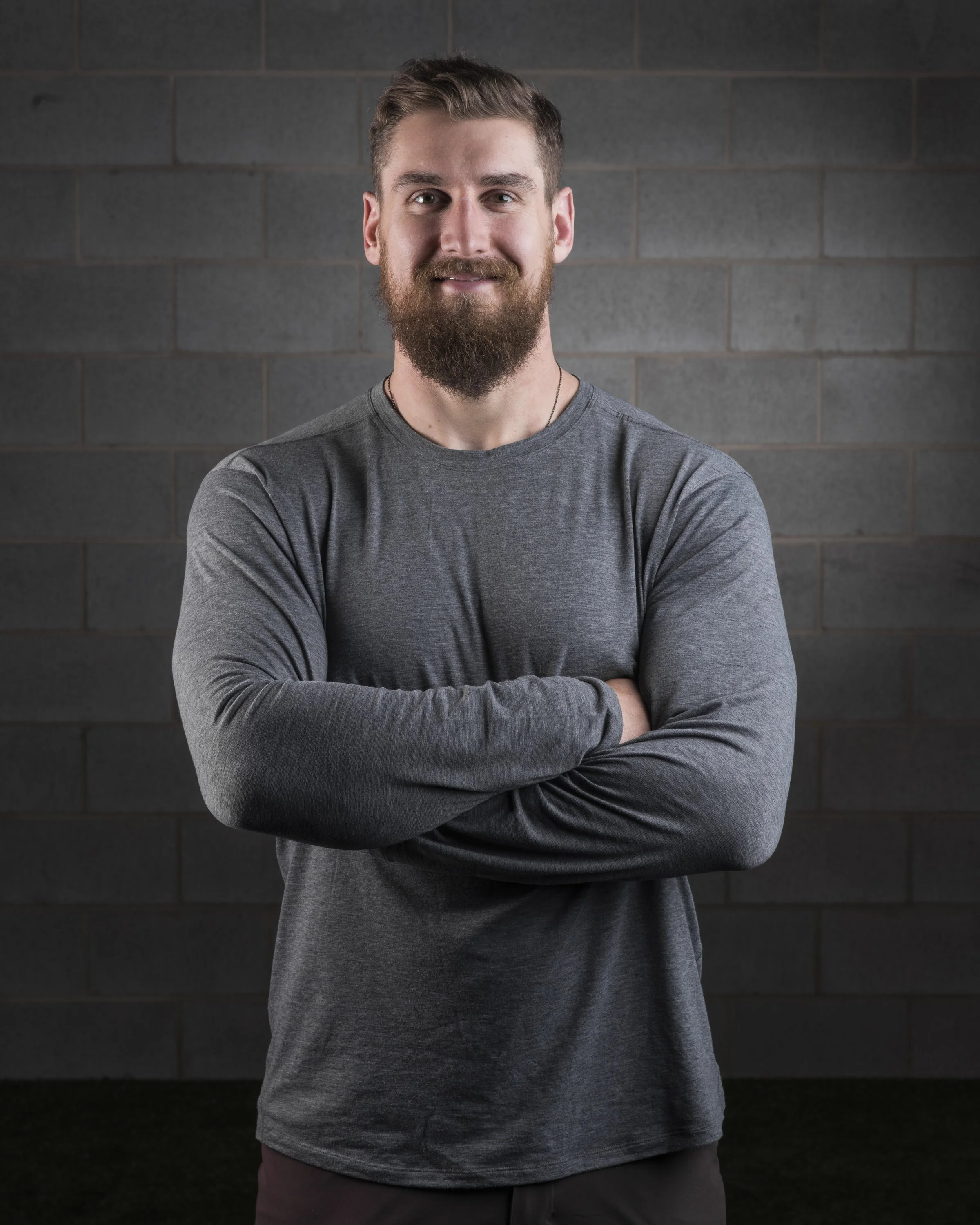 A young man with a beard and mustache, wearing a gray long-sleeve shirt, standing with arms crossed against a gray brick wall.