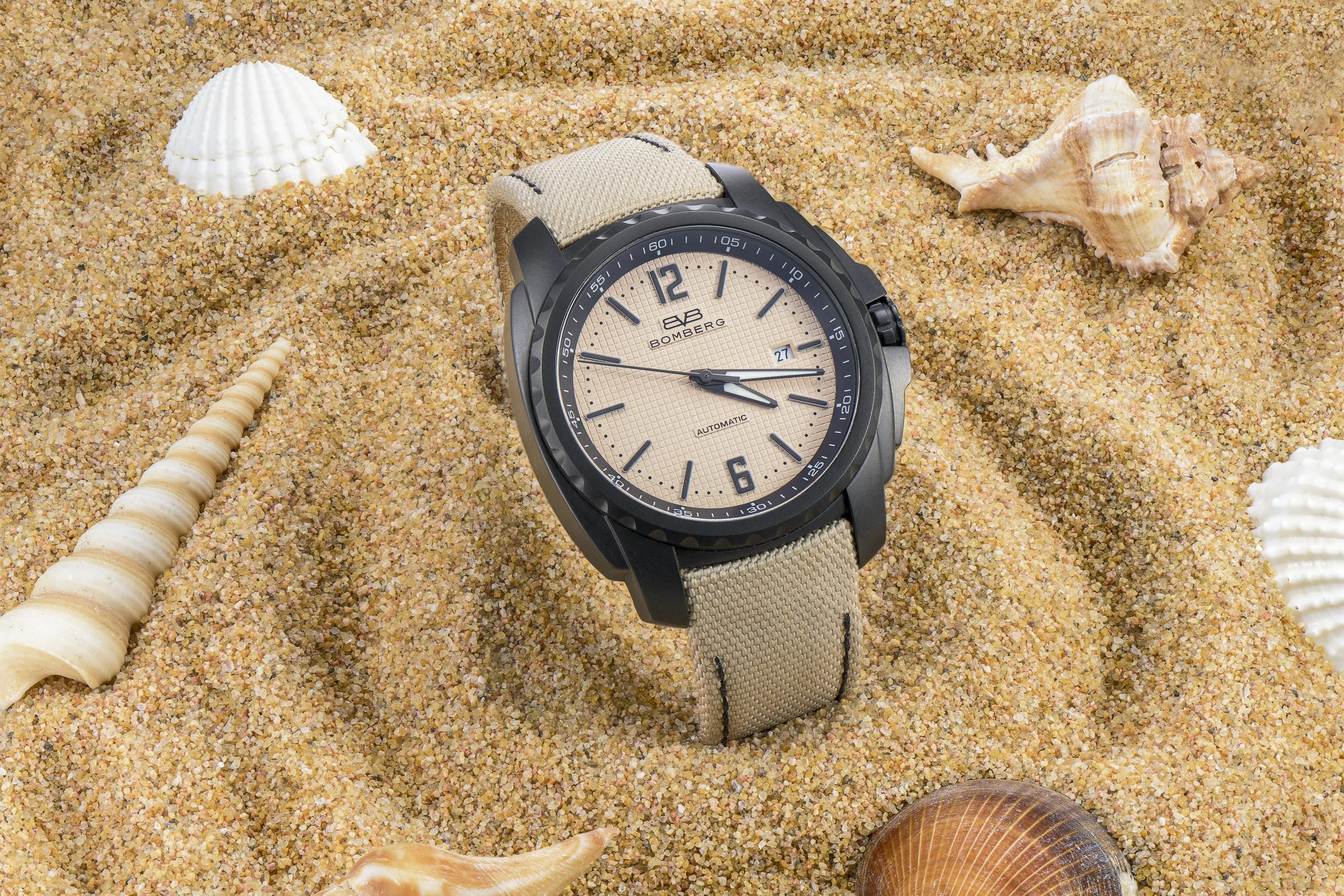 A beige and black wristwatch placed in sand surrounded by seashells on a beach.