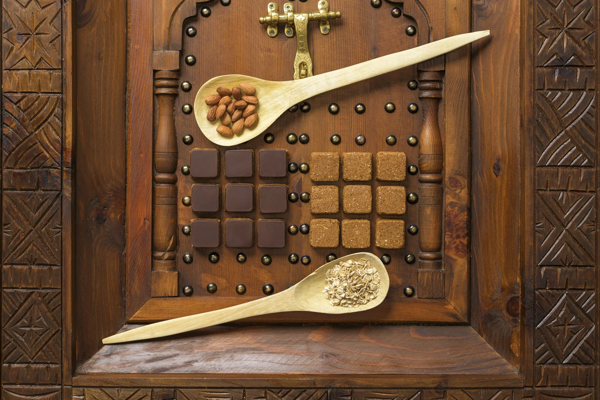 Assorted chocolates, brown sugar cubes, and rolled oats arranged on a wooden tray with carved patterns. Two wooden spoons hold almonds and oats.