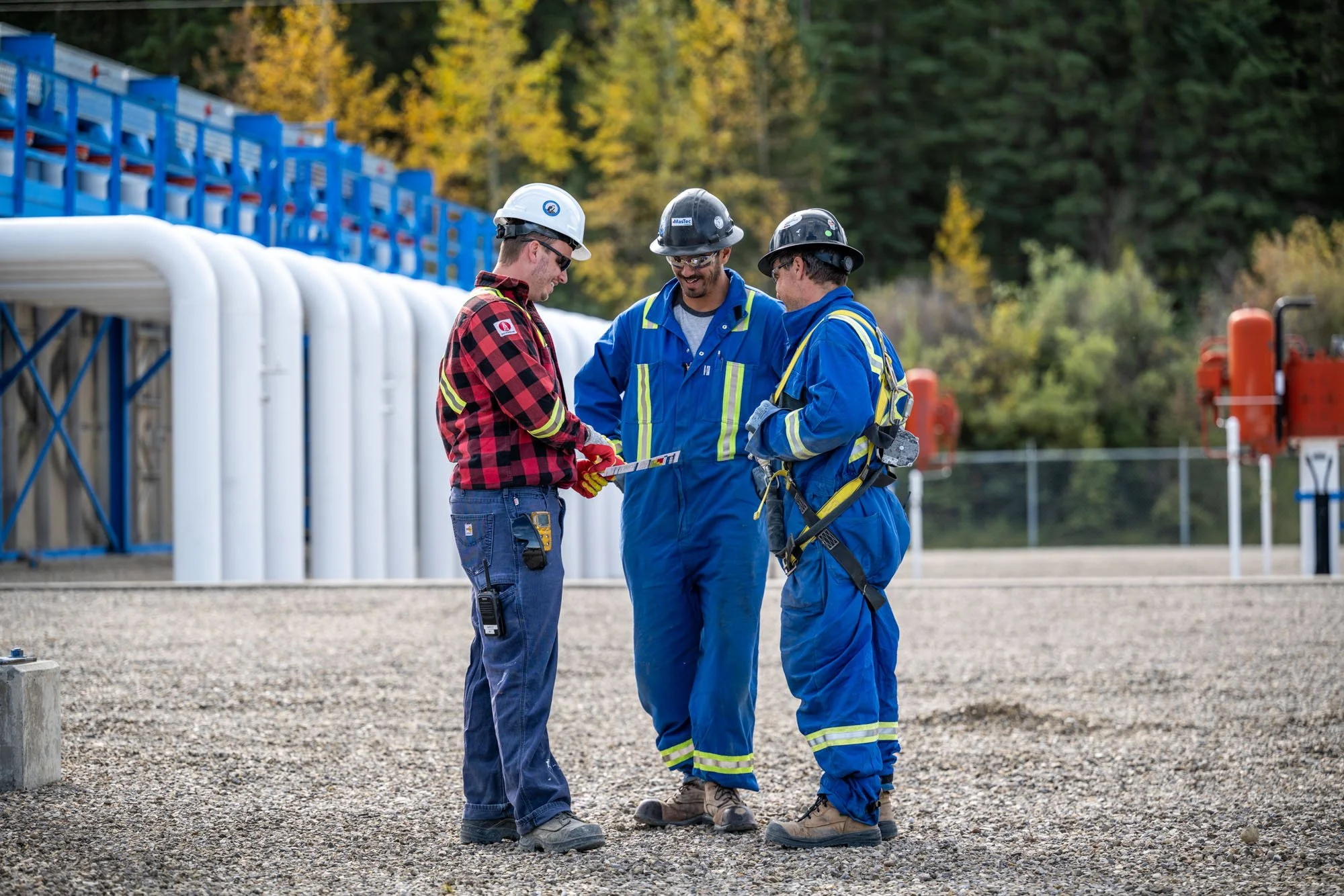 Three workers in safety gear standing outdoors, reviewing plans at a construction site with trees and equipment in the background.