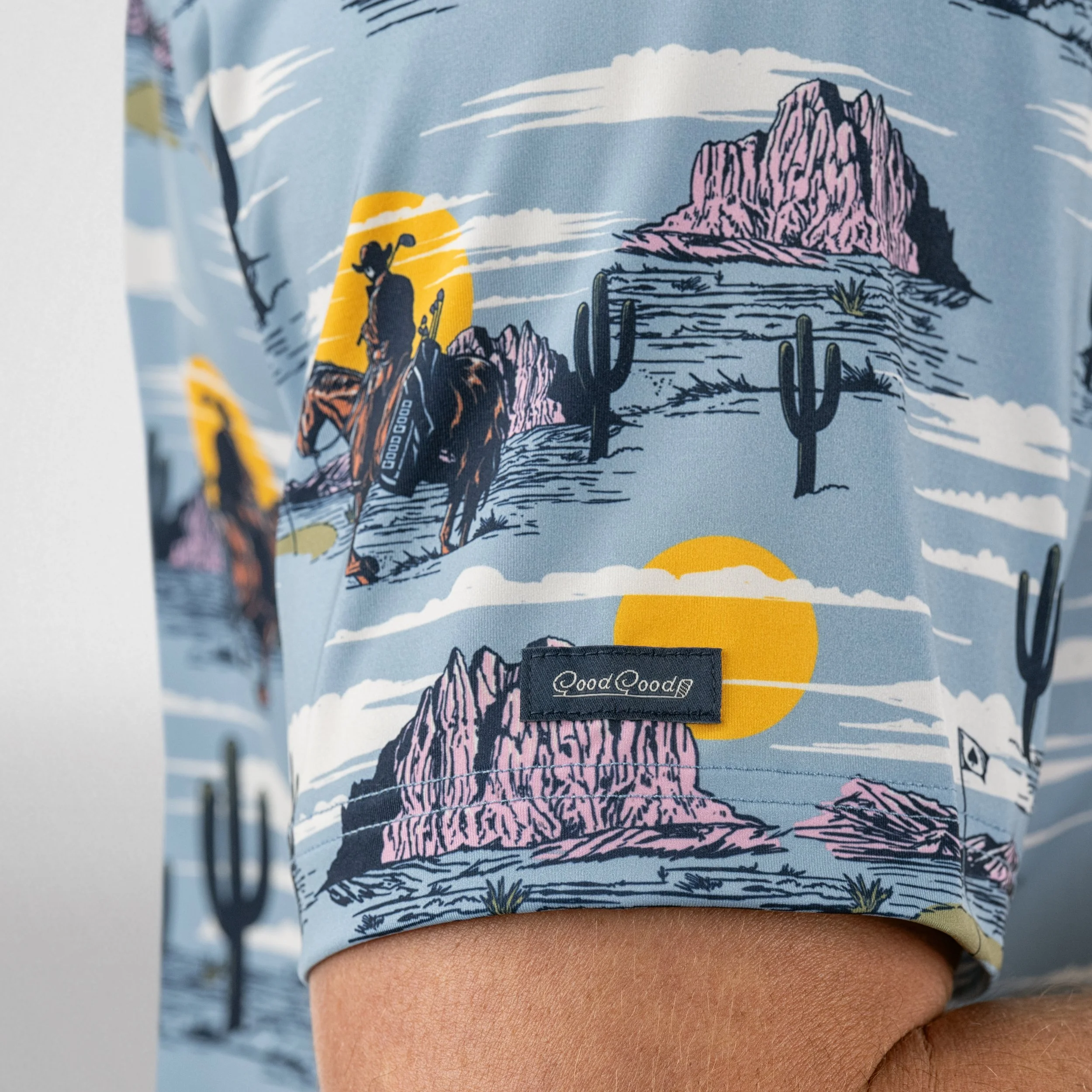 Close-up of a fabric with a desert scene featuring pink mountains, cacti, a cowboy on horseback, and a setting sun, with a label reading 'Good Good' on the sleeve.