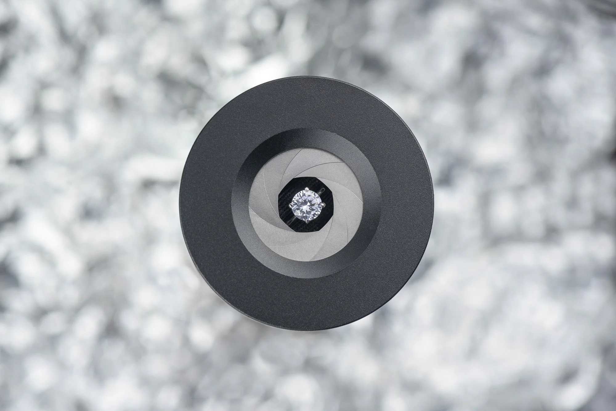 Close-up of a camera lens with a diamond in the center, against a blurry, metallic, and shiny background.