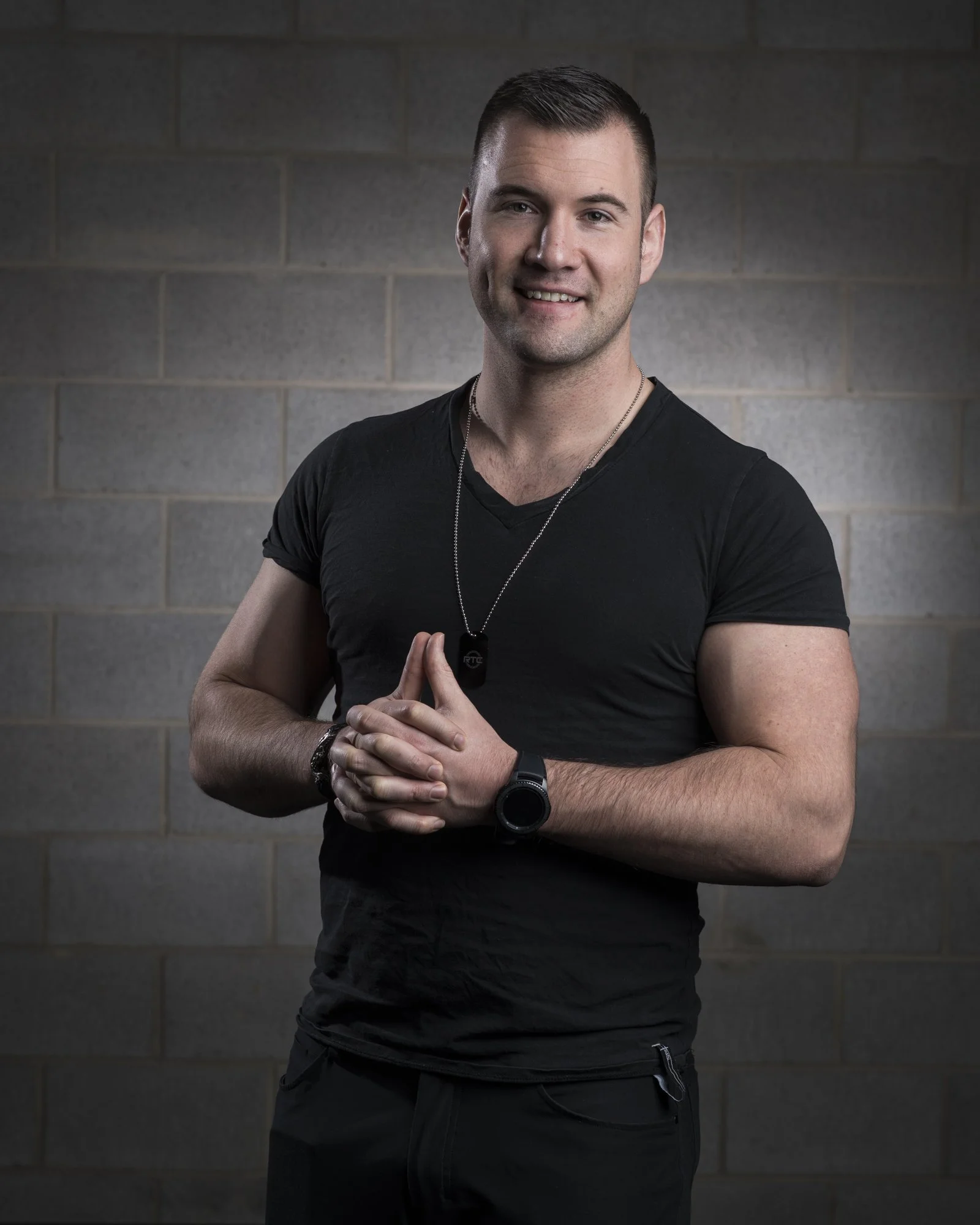 A smiling man standing with hands clasped in front of a gray brick wall, wearing a black v-neck t-shirt, a wristwatch, a necklace, and a dog tag.
