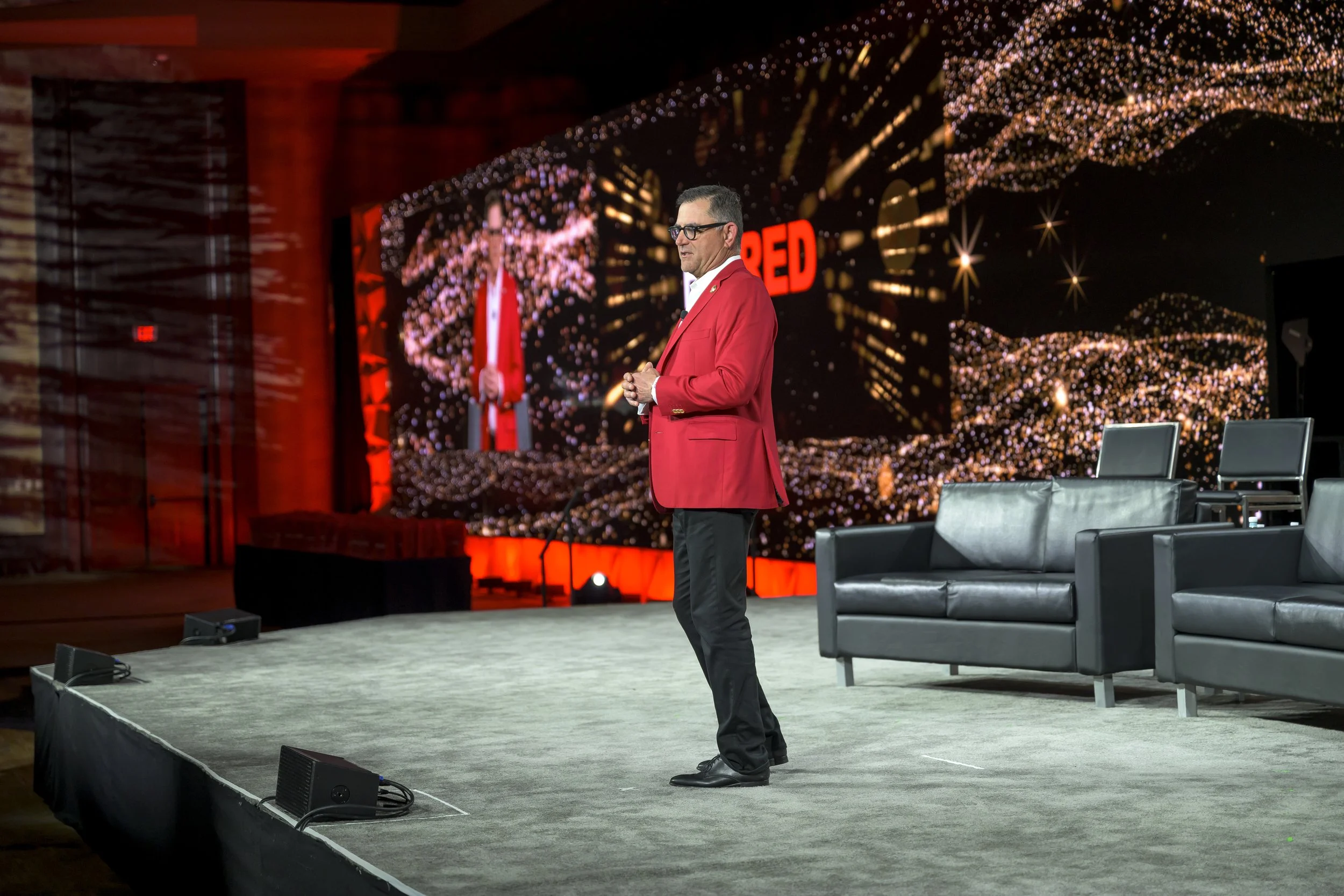 A man in a red blazer and black pants speaking on stage at a conference, with large screens behind him displaying the word 'RED' and swirling golden lights.
