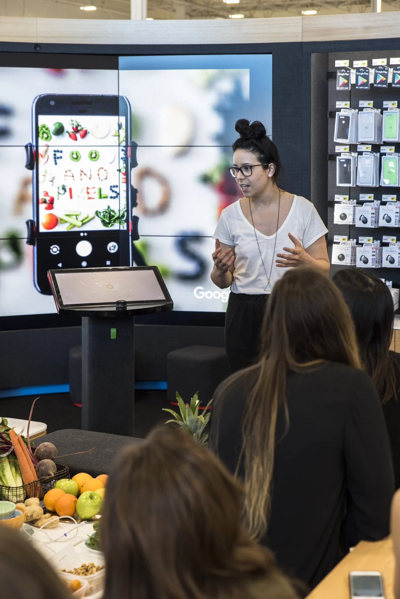 A woman giving a presentation to a small group in a retail store, with a large digital screen behind her showing an image of a smartphone with food items and text.