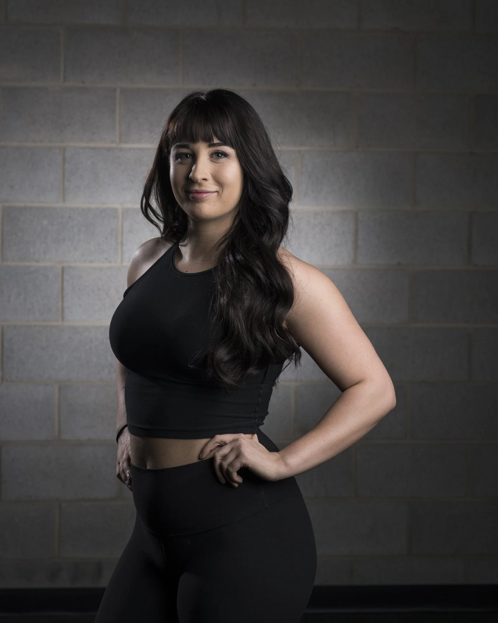 A woman in black athletic wear standing against a gray concrete block wall.