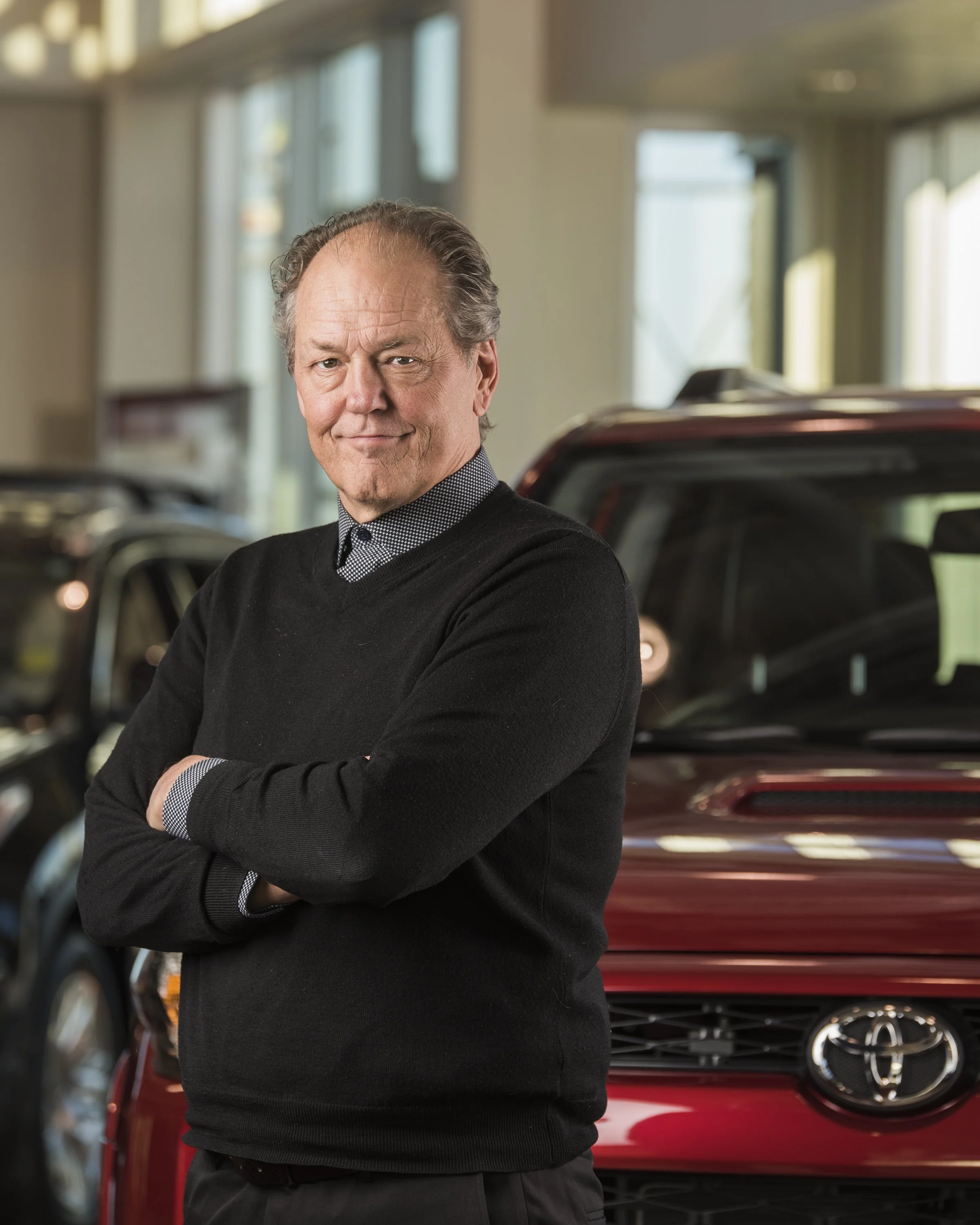 A man standing with crossed arms in a car dealership filled with Toyota vehicles, including a red SUV behind him.