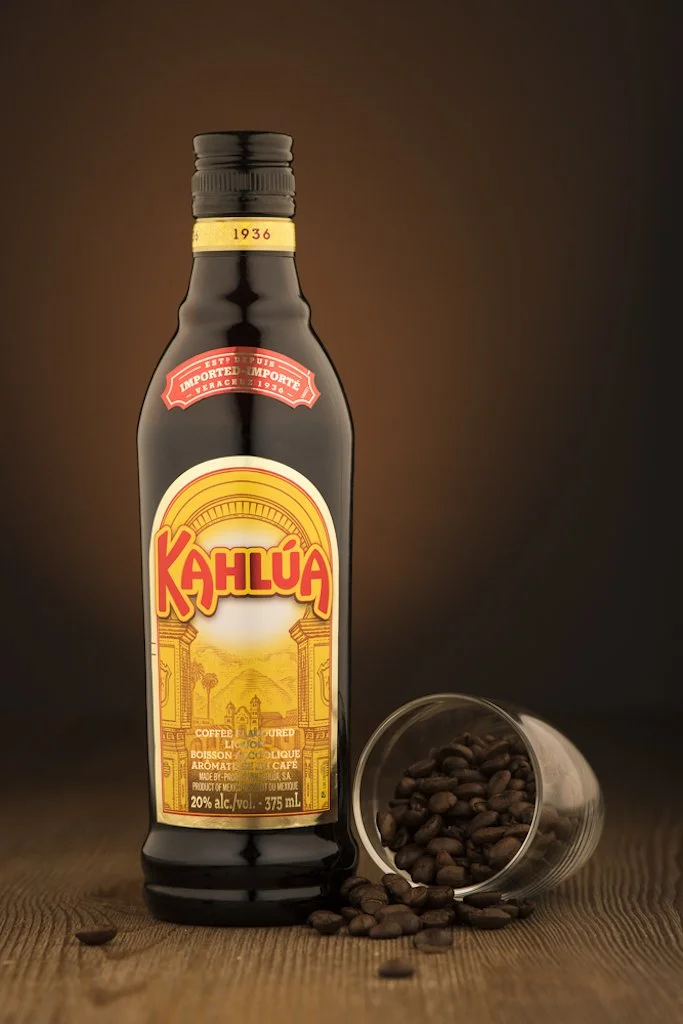 Kahlua coffee liqueur bottle with a glass tipped over and spilling coffee beans on a wooden surface.