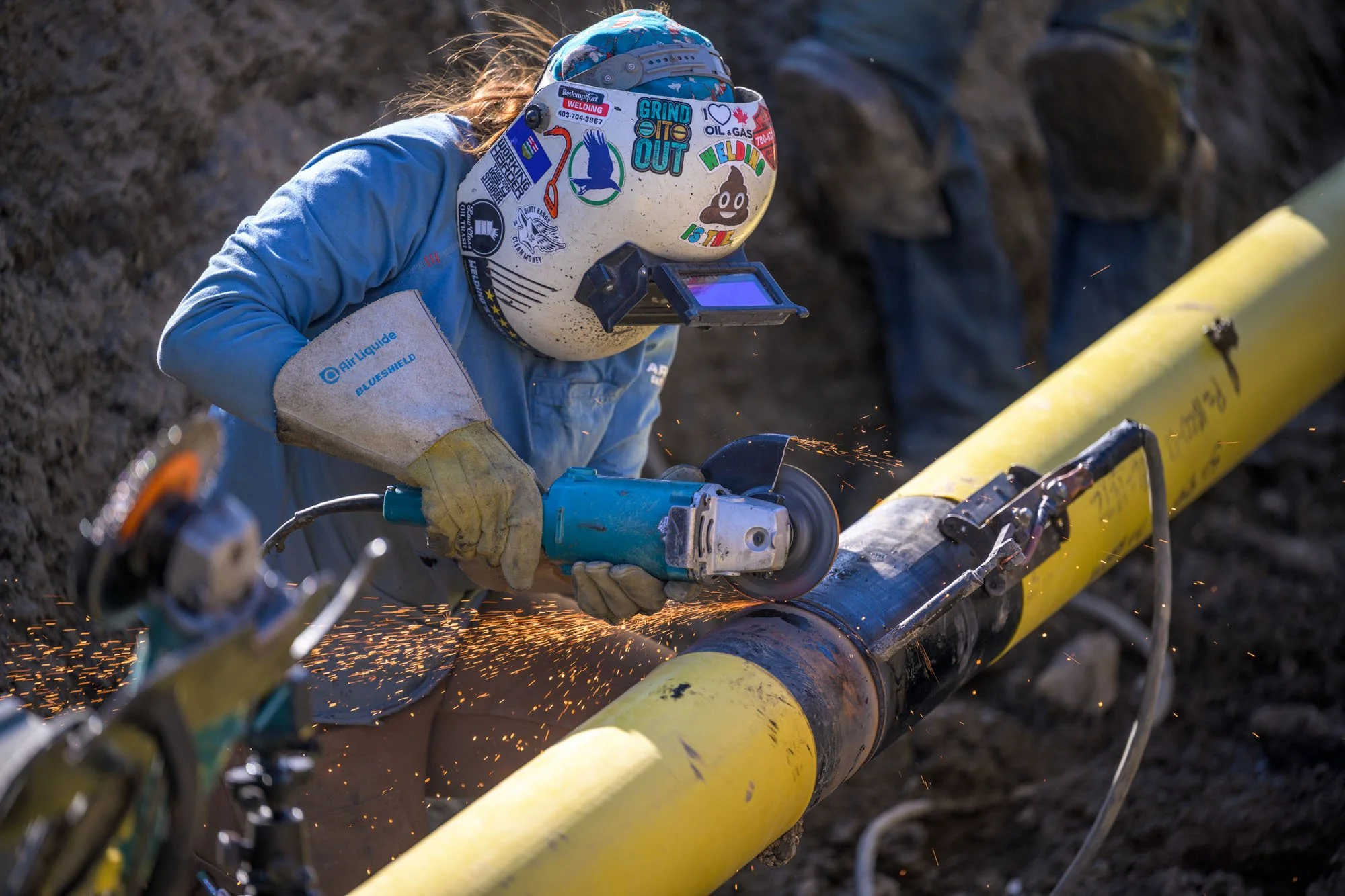 A worker welding a yellow pipeline outdoors. The worker is wearing a protective helmet with various stickers, a blue long-sleeved shirt, and gloves while using a power tool that generates sparks.