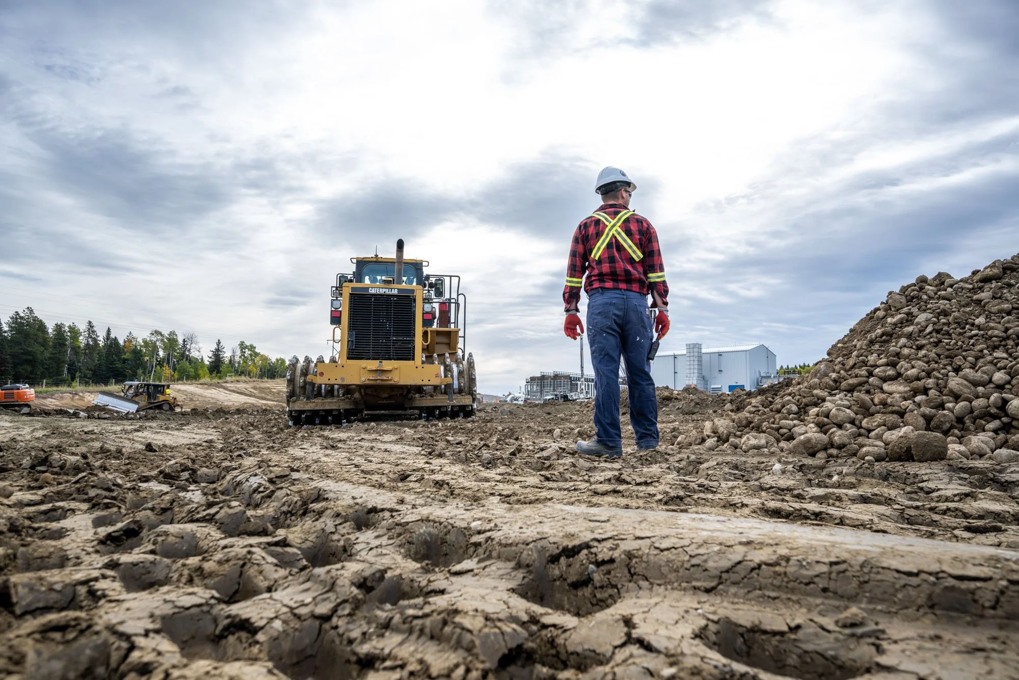 Construction worker in safety gear standing on dirt with large pile of rocks and construction machinery in background.