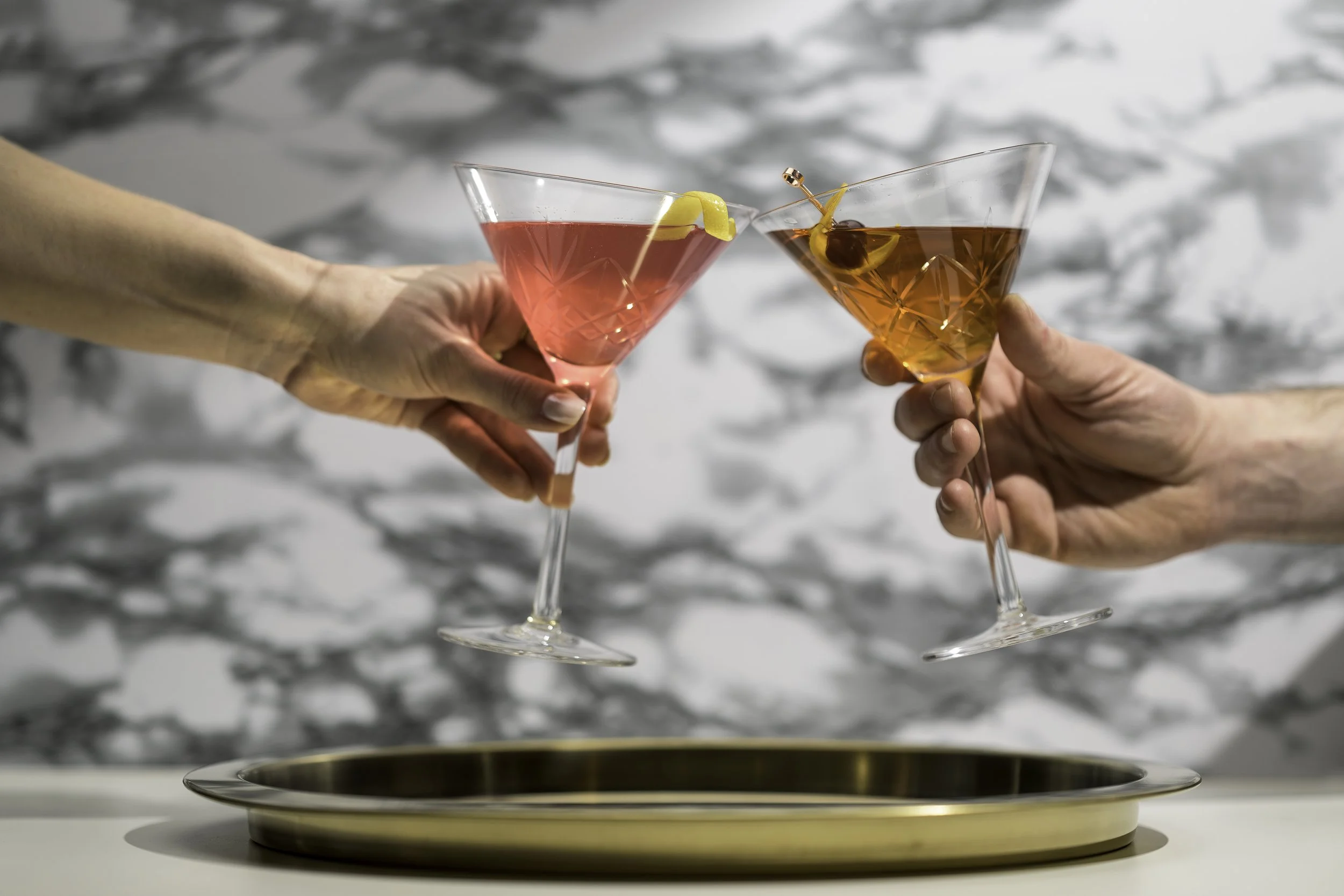 Two people clink martini glasses filled with pink and orange cocktails, garnished with lemon and cherry, over a gold tray on a white surface with a marble pattern background.