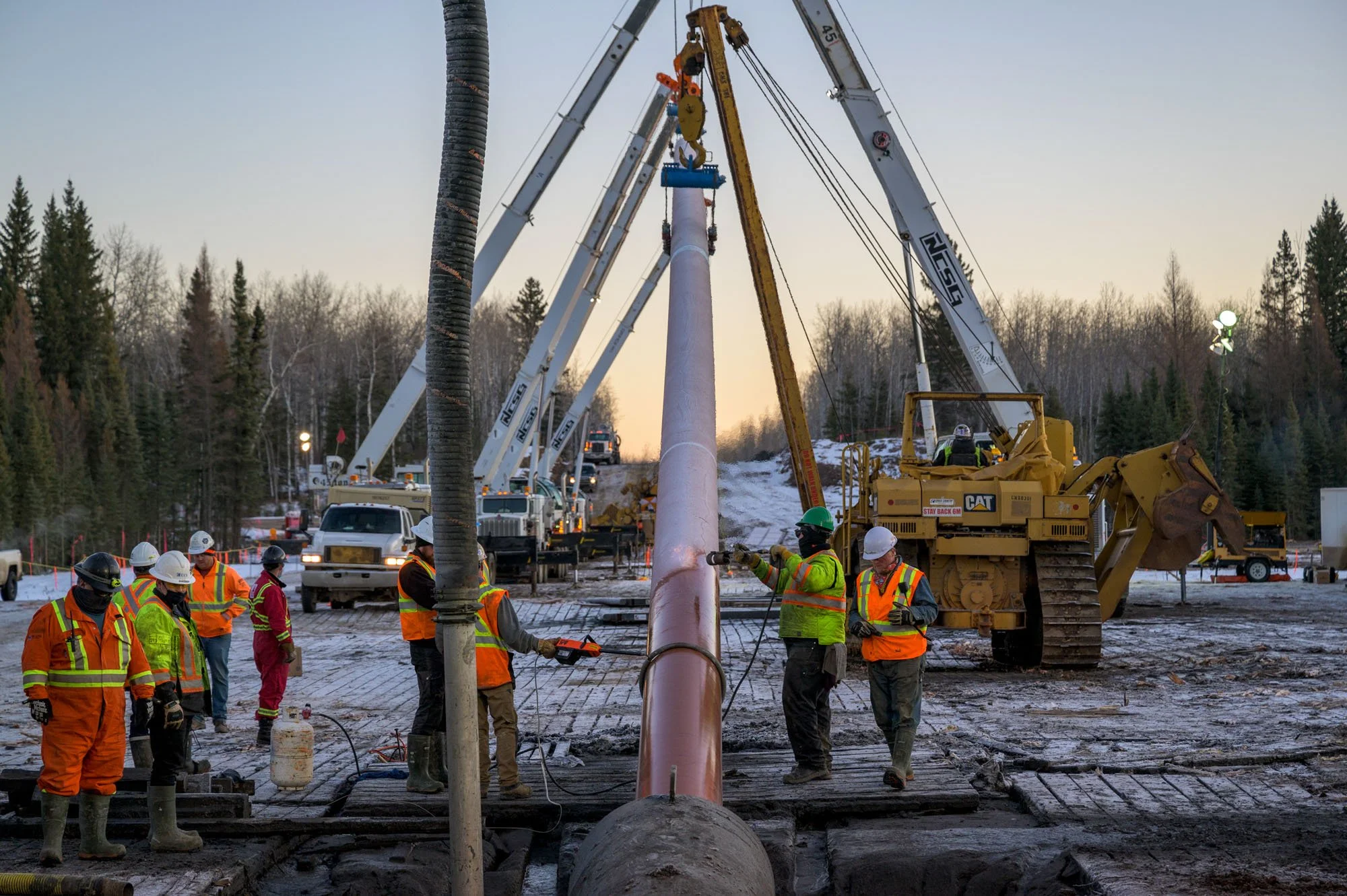 Construction workers in safety gear working with large machinery to install or repair a large pipe at a construction site in a wooded area during sunset.