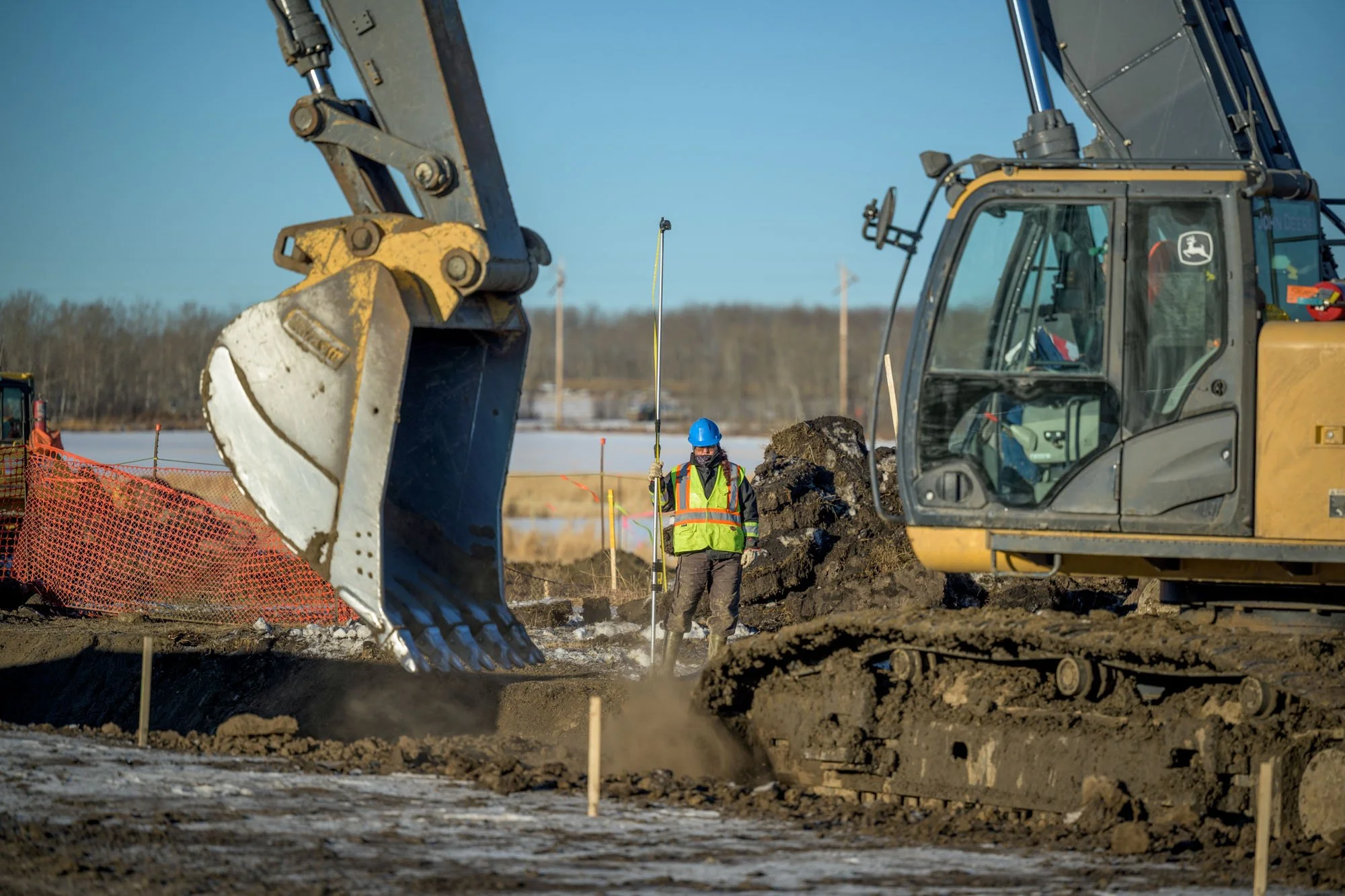 A construction worker wearing a blue helmet and reflective vest standing between large construction machinery on a dirt site, with muddy ground and a partially cloudy sky in the background.