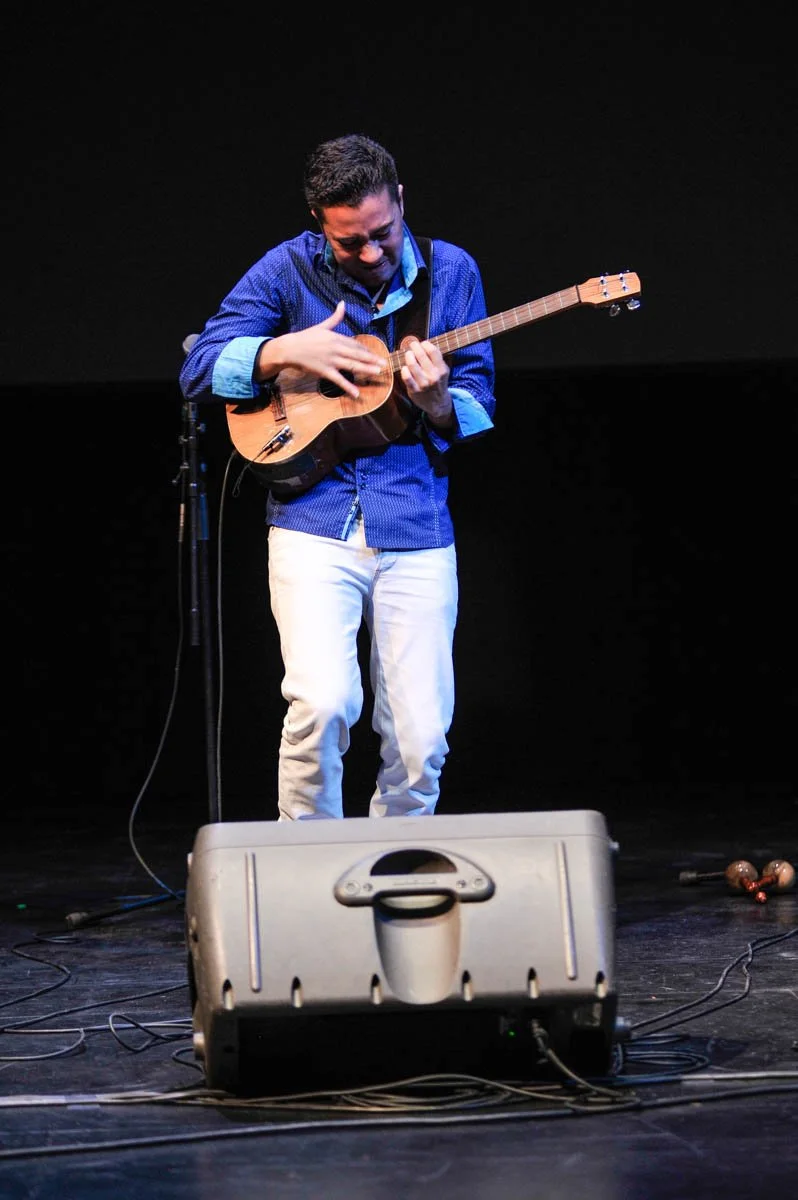 A man on stage playing a ukulele, wearing a blue shirt and white pants, with a black background and stage equipment in view.