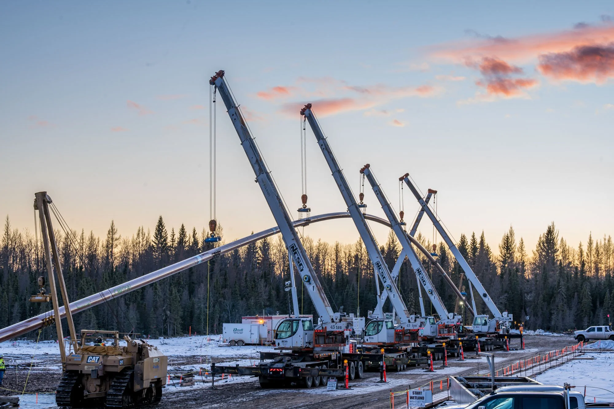 Construction site with multiple cranes lifting a long metal pipe or pole, set against a background of a forest and a sunset sky.