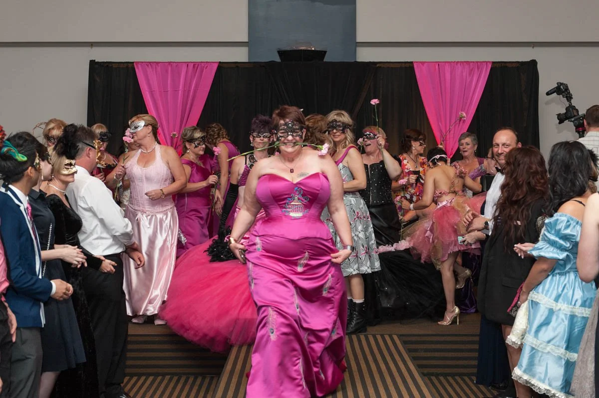 Women wearing elaborate pink gowns and masks at a costume party or event.