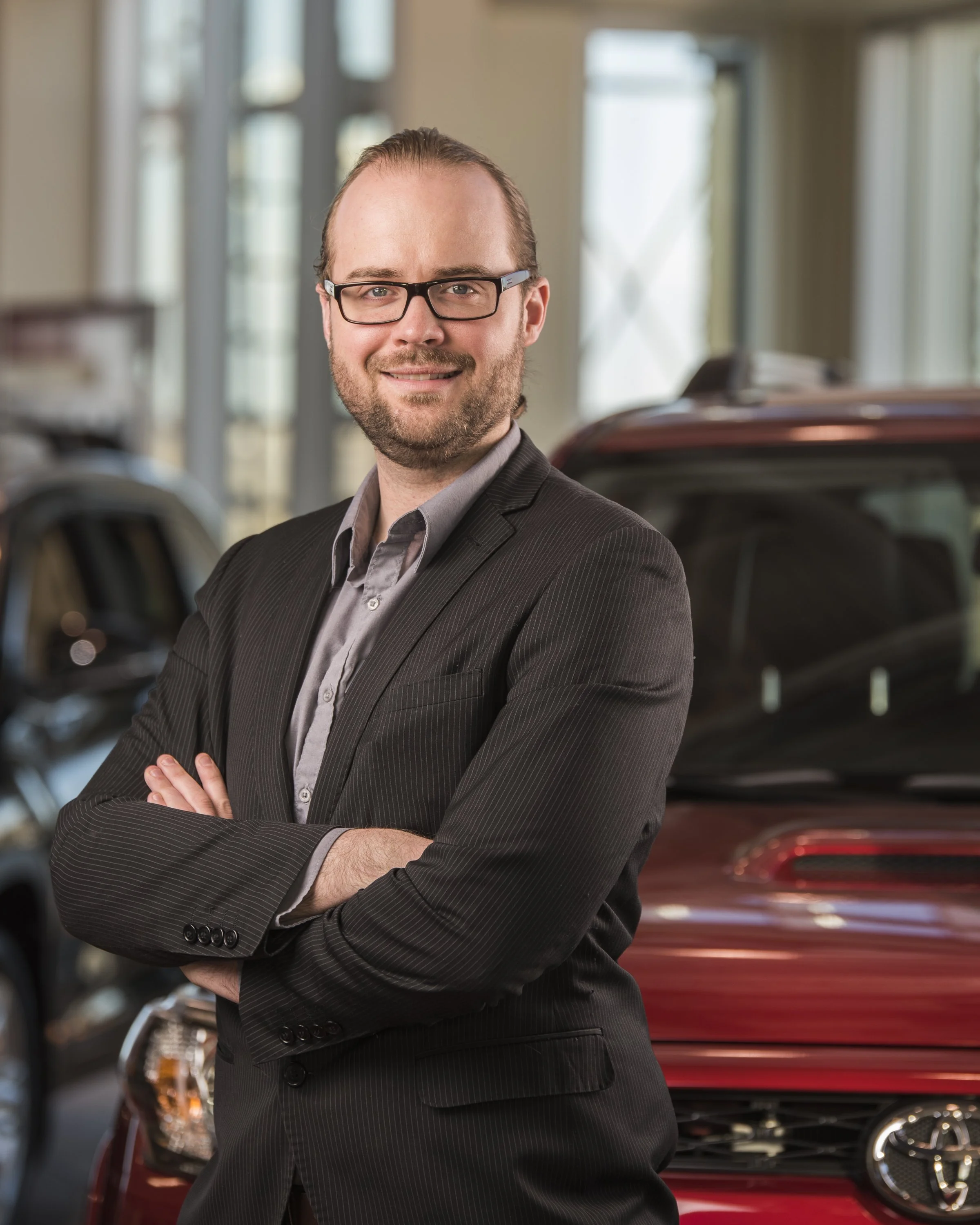 A man wearing glasses and a dark suit with a gray shirt standing with arms crossed in a car dealership. There are cars visible in the background.