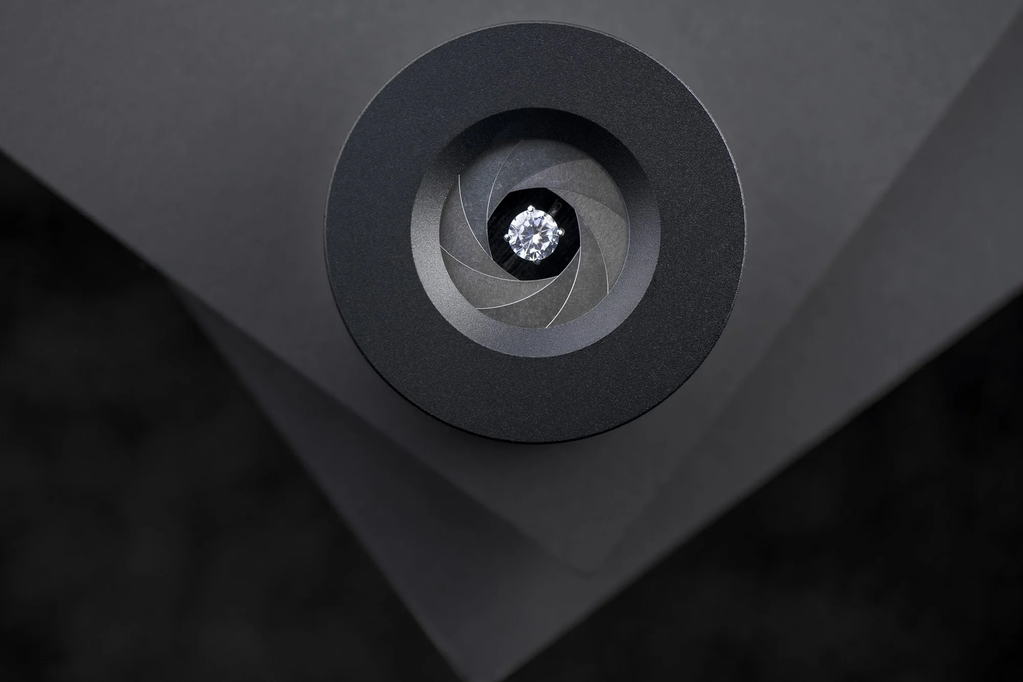 Close-up of a black camera lens with a diamond-like reflection at the center, attached to a dark gray device with angular edges.