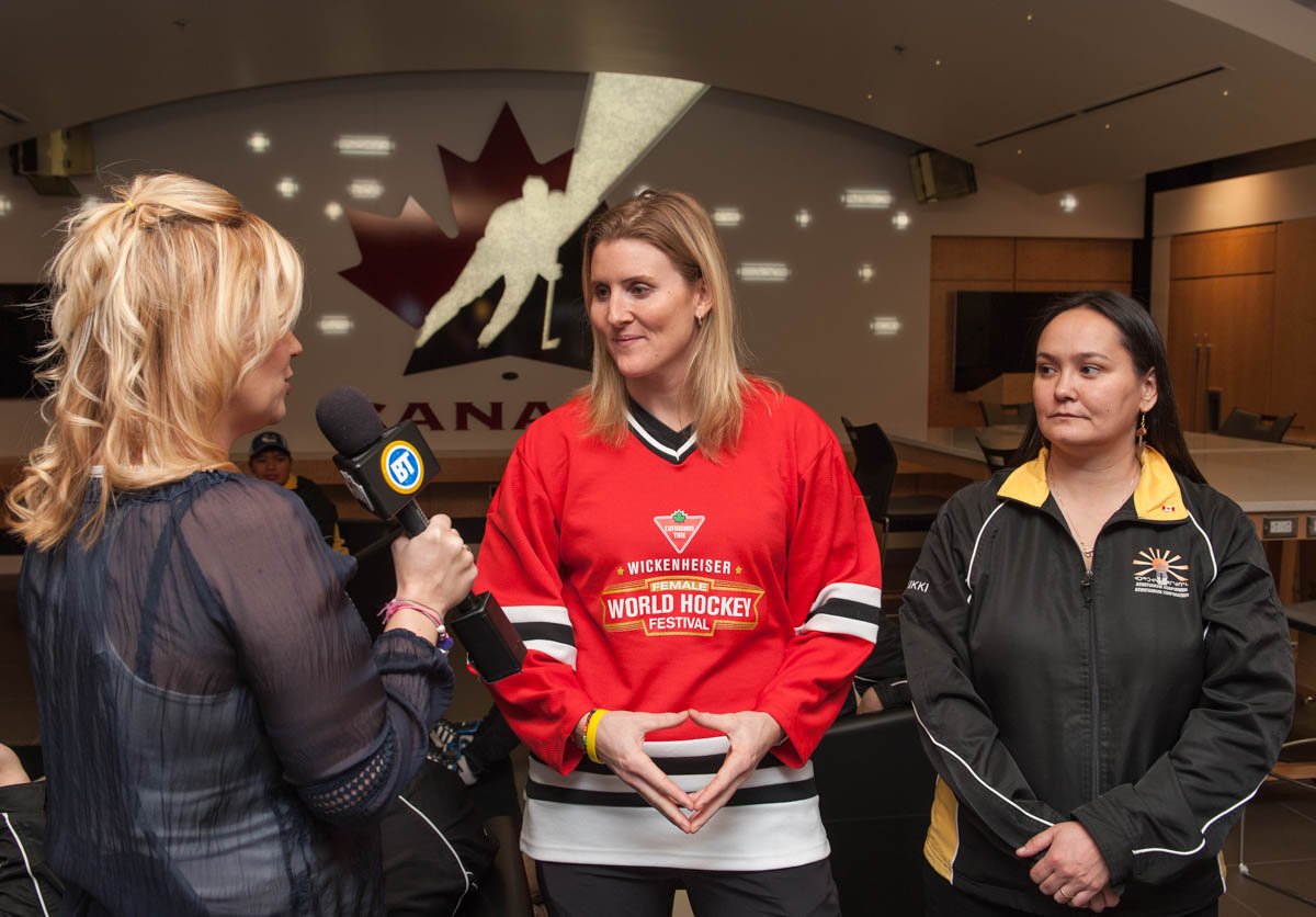 A woman with blond hair interviewing two women with dark hair in a room with a large logo of Canada and a hockey player on the wall.
