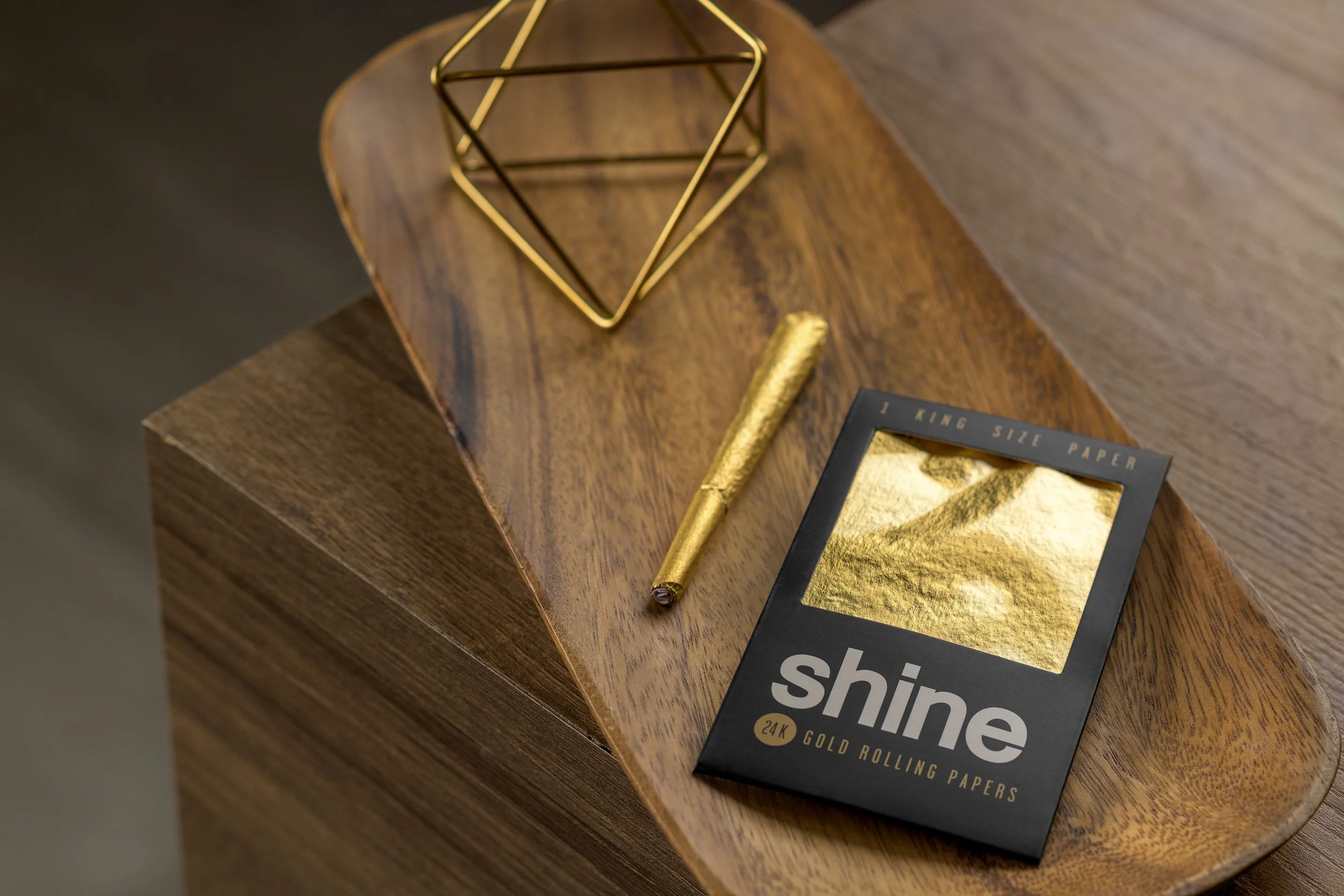 A wooden tabletop with black box of 24K gold rolling papers, a gold pen, and a geometric gold wire decorative object.