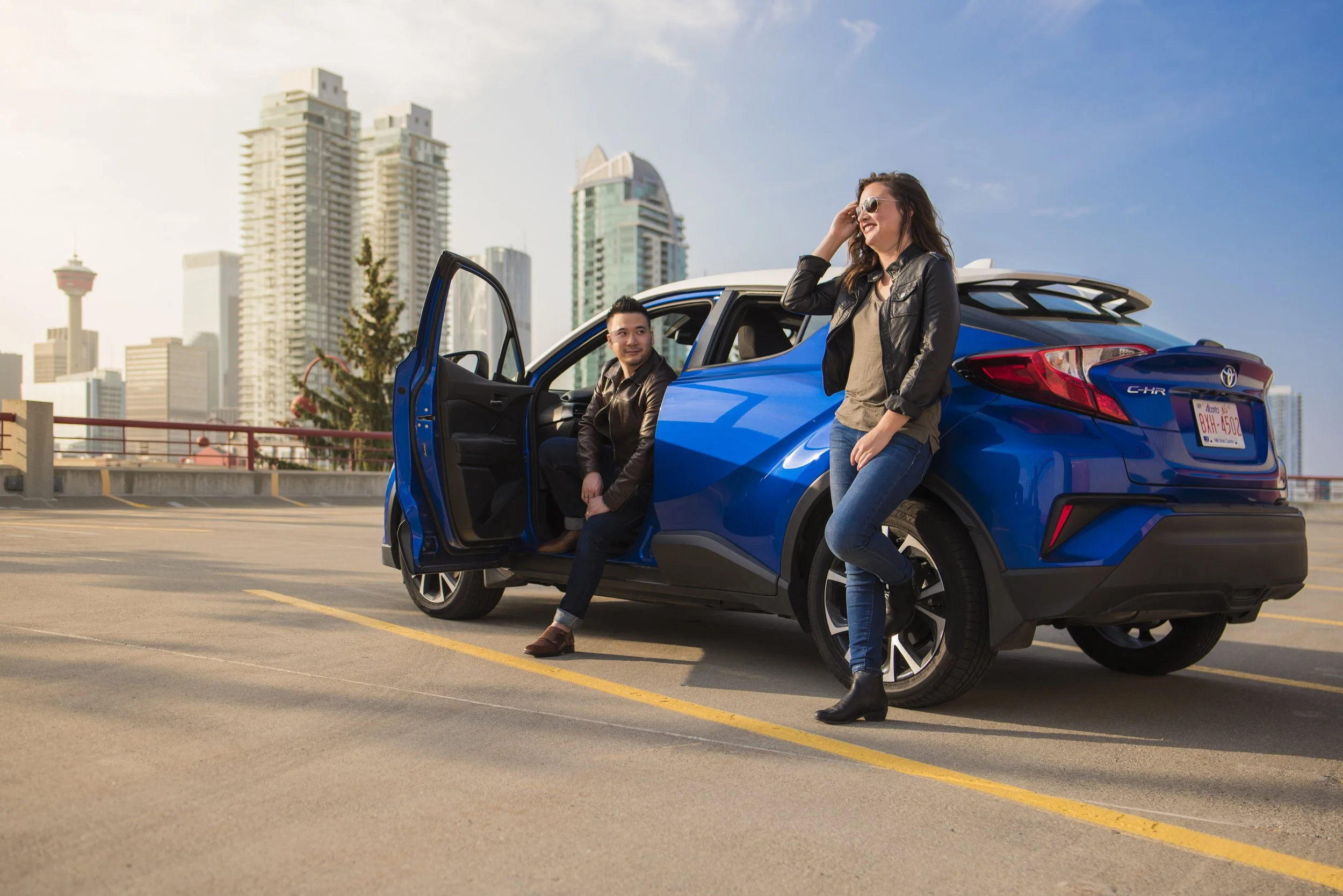 A man and woman are posing with a blue Toyota C-HR in a parking lot with a city skyline in the background. The man is sitting in the car, and the woman is standing next to it, smiling and adjusting her sunglasses.