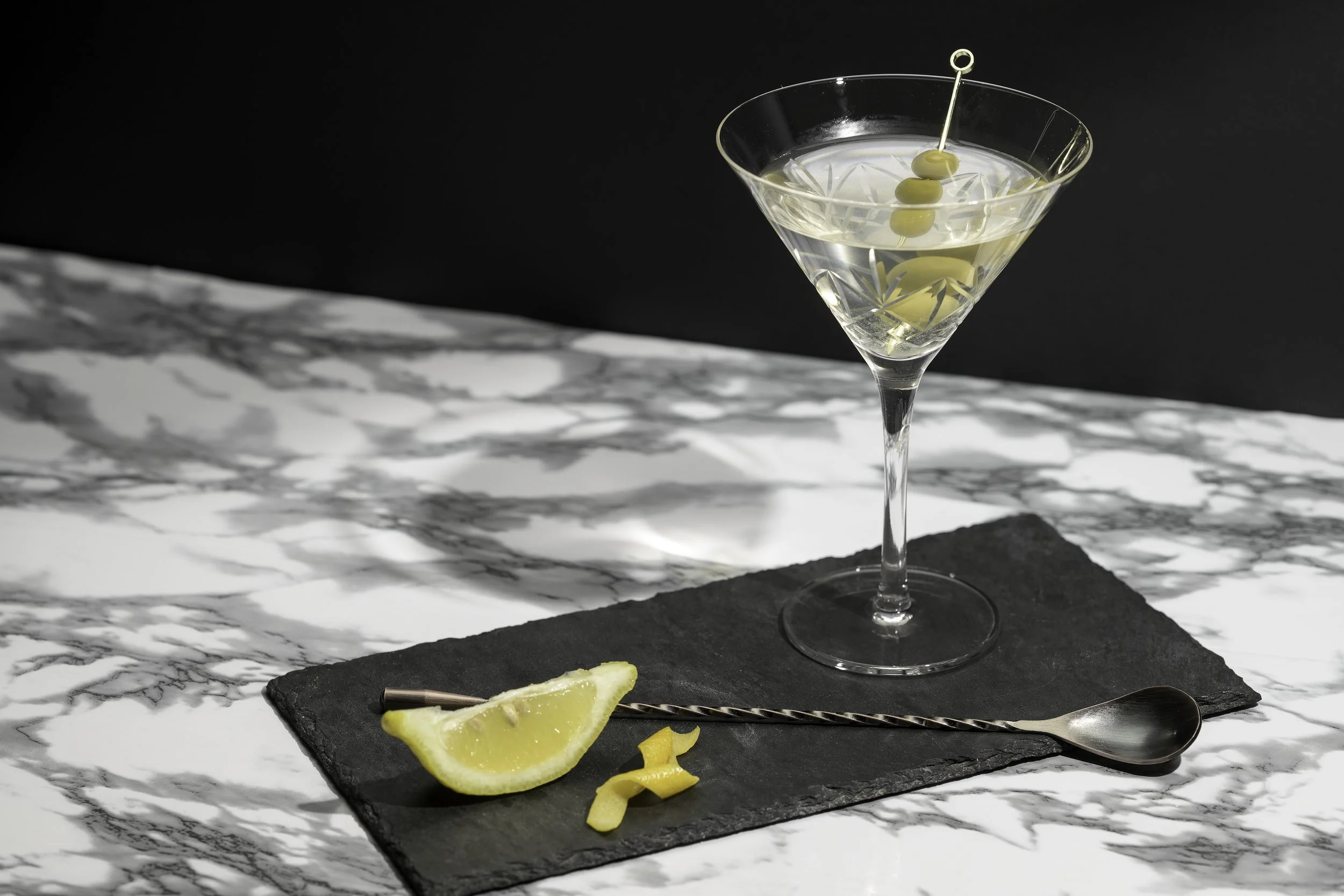 A cocktail glass with a clear drink and olive skewers, a lemon wedge, and lemon peel on a black slate serving board on a marble countertop.