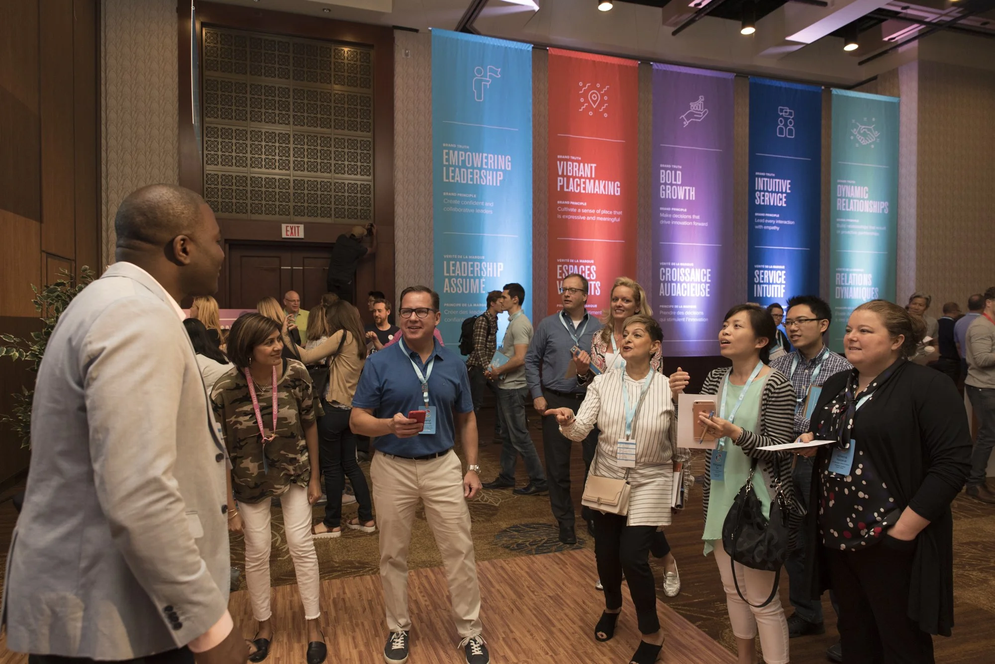 A group of diverse people in business casual attire engaging in conversation at a conference or seminar. The background features large colorful banners with leadership and growth themes.