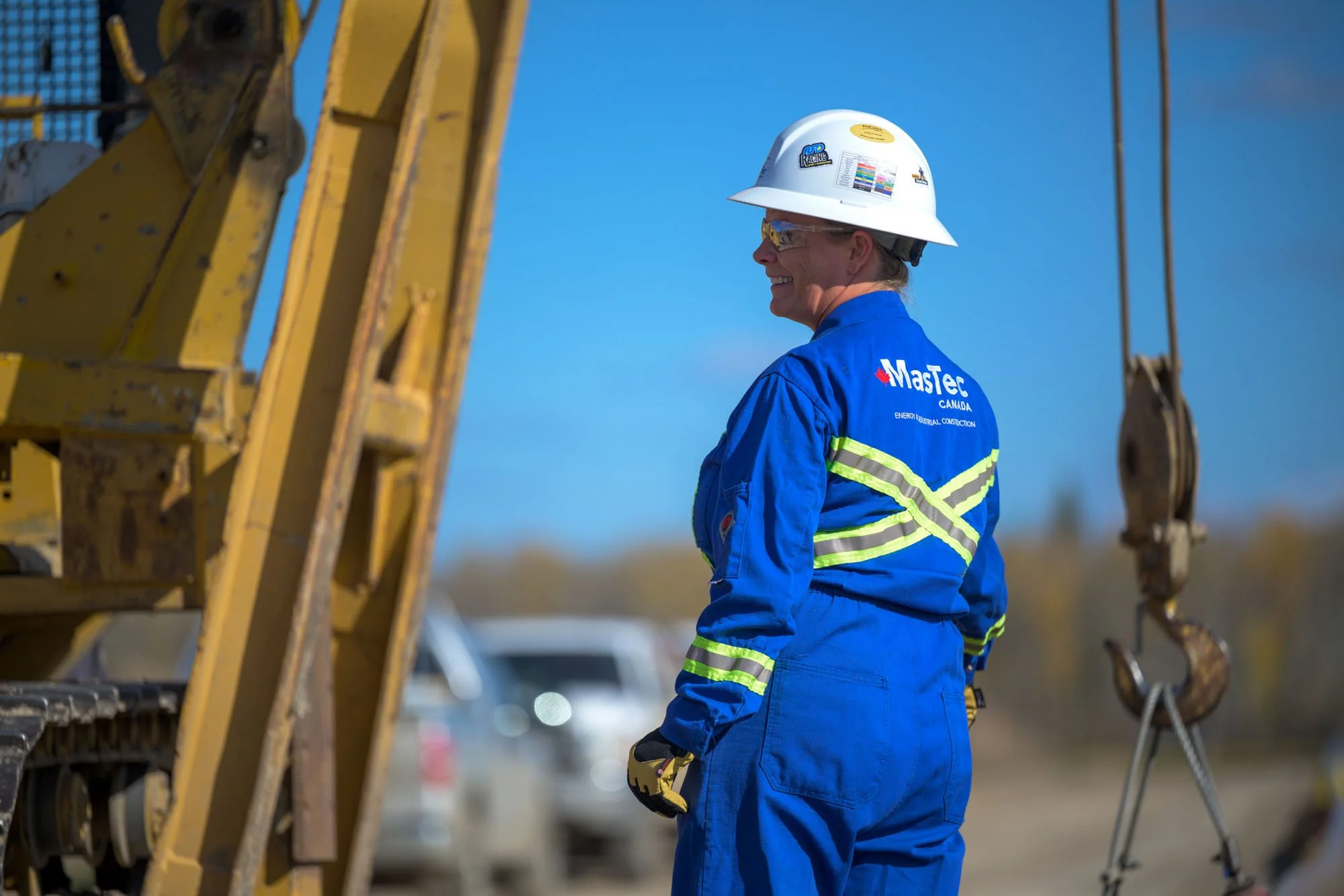 A woman in blue safety gear and a white hard hat at a construction site, standing beside construction equipment with a hook.